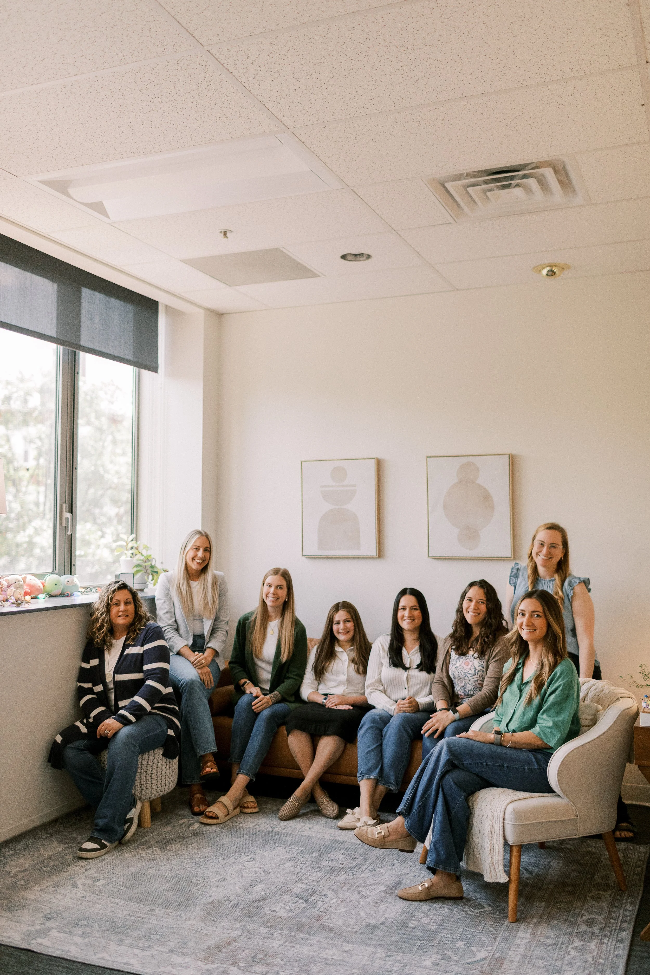 A group of women sitting and posing for a photo in a brightly lit room with art on the walls and a window with blinds nearby.