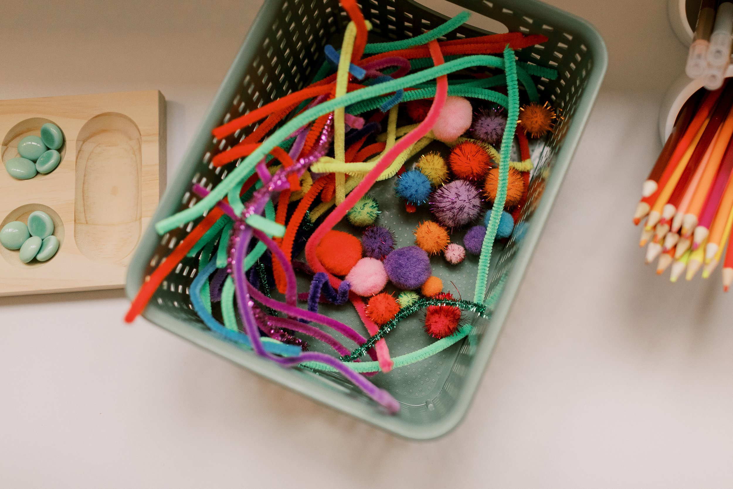 A rectangular green basket filled with colorful pipe cleaners and fuzzy pom-poms, placed on a white surface. To the side, there is a light-colored wooden tray with mint green oval candies and a slot for a small object. To the right of the basket, there is a container holding yellow and orange pencils with erasers.