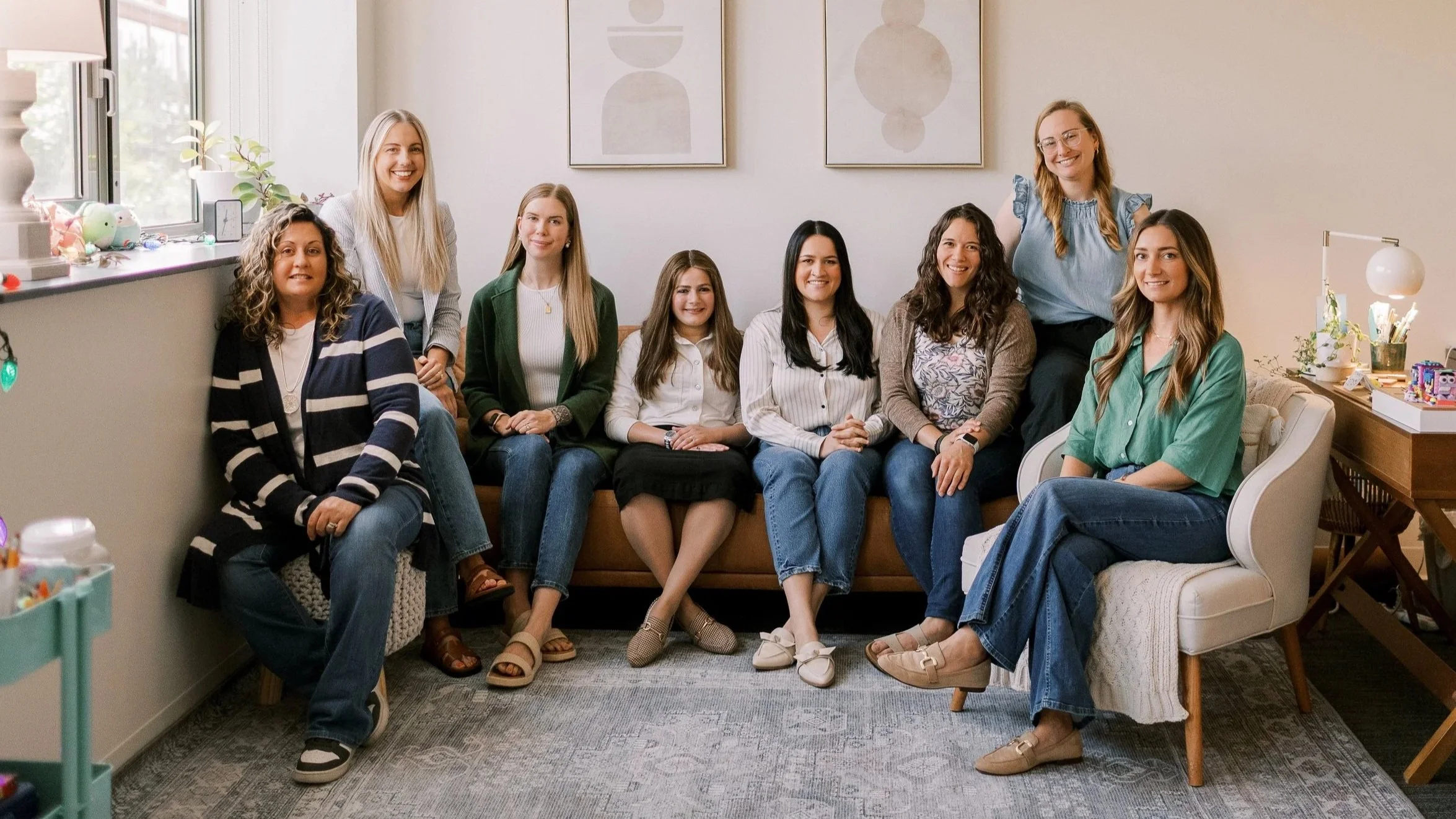 A group of nine women sitting and standing in a cozy living room with minimalistic art on the wall, plants, and decorated shelves, smiling at the camera.