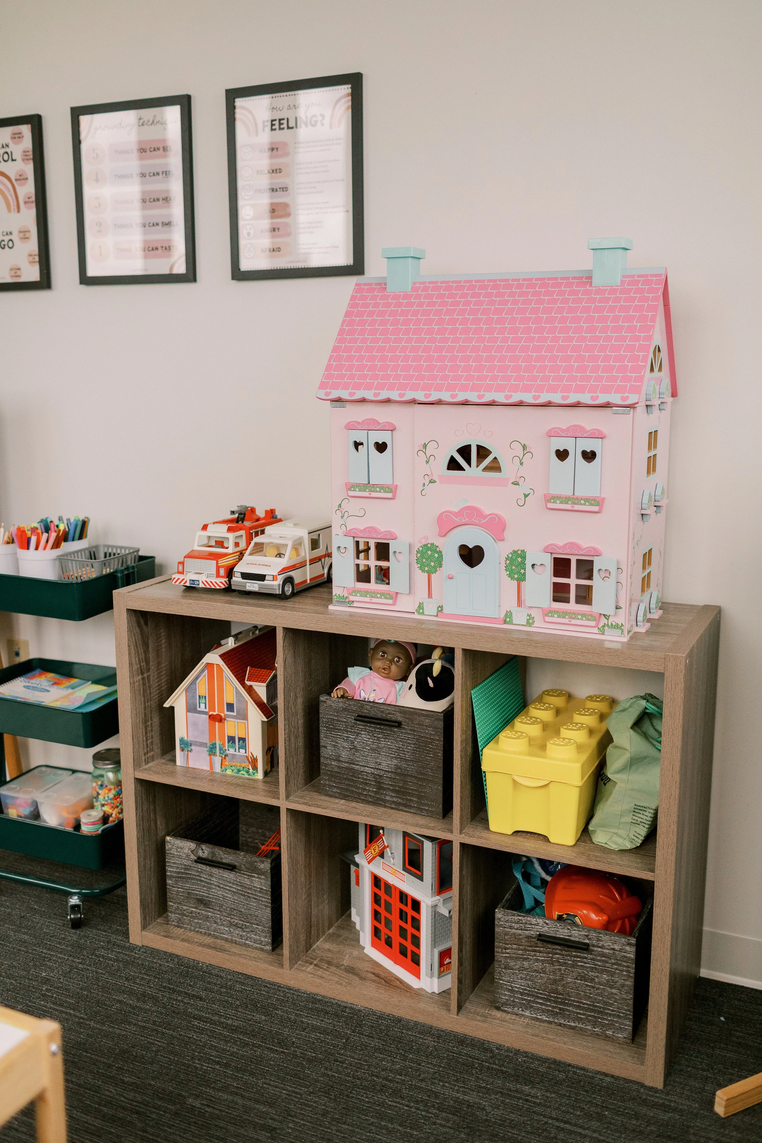 A wooden shelf with compartments containing toys, including a doll, a toy house, a book, and various storage boxes. On top of the shelf is a pink dollhouse, and behind it, a wall with framed posters about feelings and emotions.