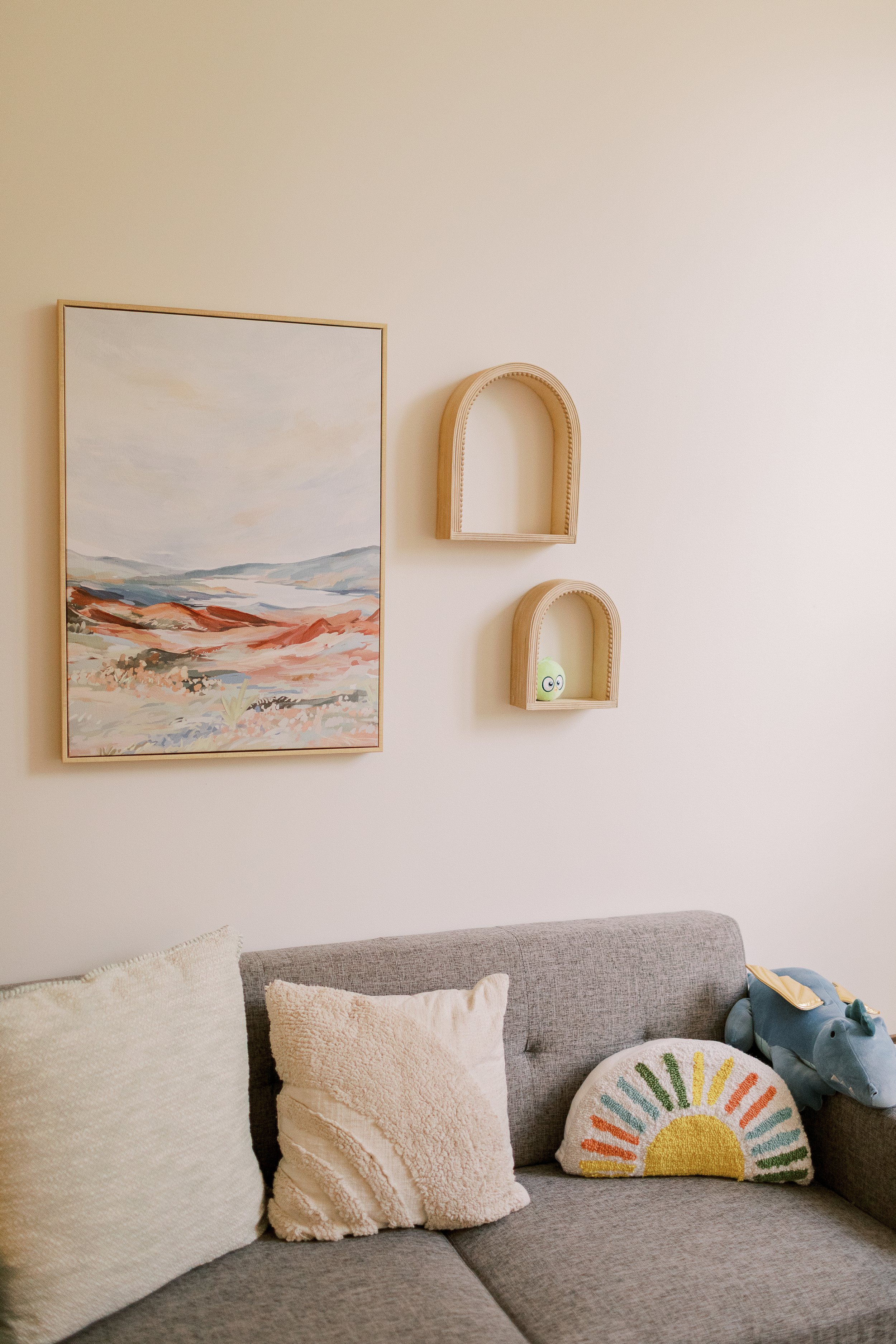 Living room with beige wall, watercolor landscape painting, and two wooden half-circle shelves. Cushions and stuffed animal on gray sofa, rainbow-patterned pillow