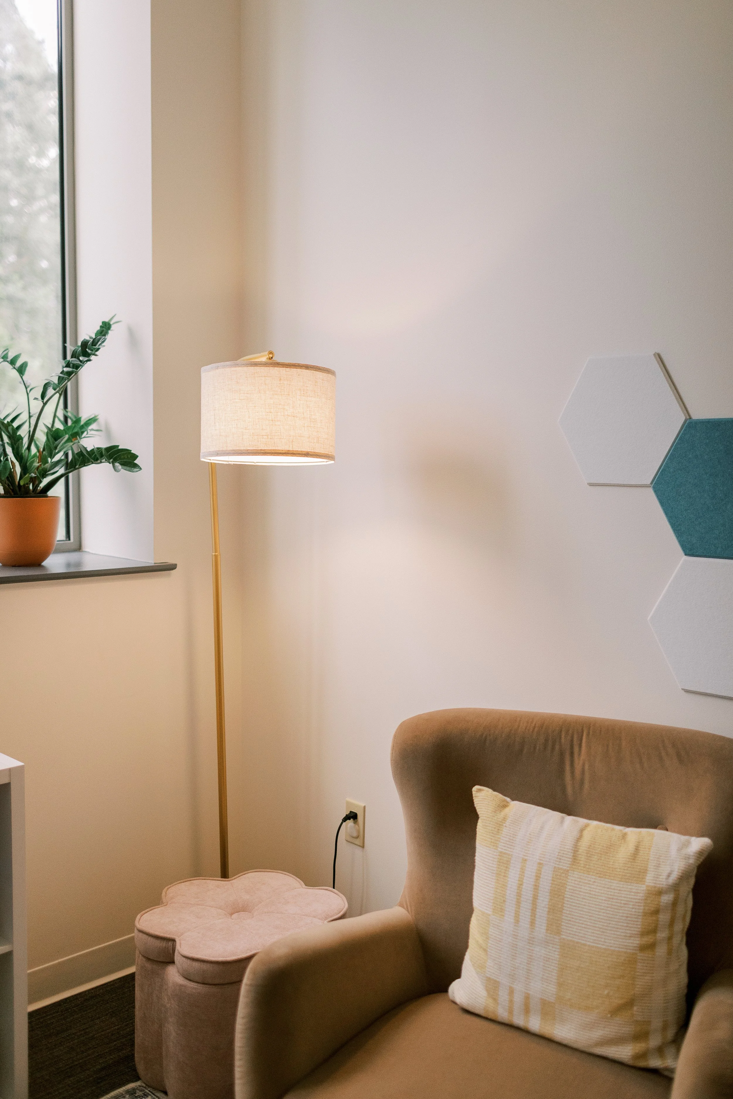 A cozy living room corner with a beige armchair and a matching pink ottoman, a pillow with yellow and white checkered pattern, a potted plant on the windowsill, a standing lamp, and three decorative hexagon wall panels in white and blue.