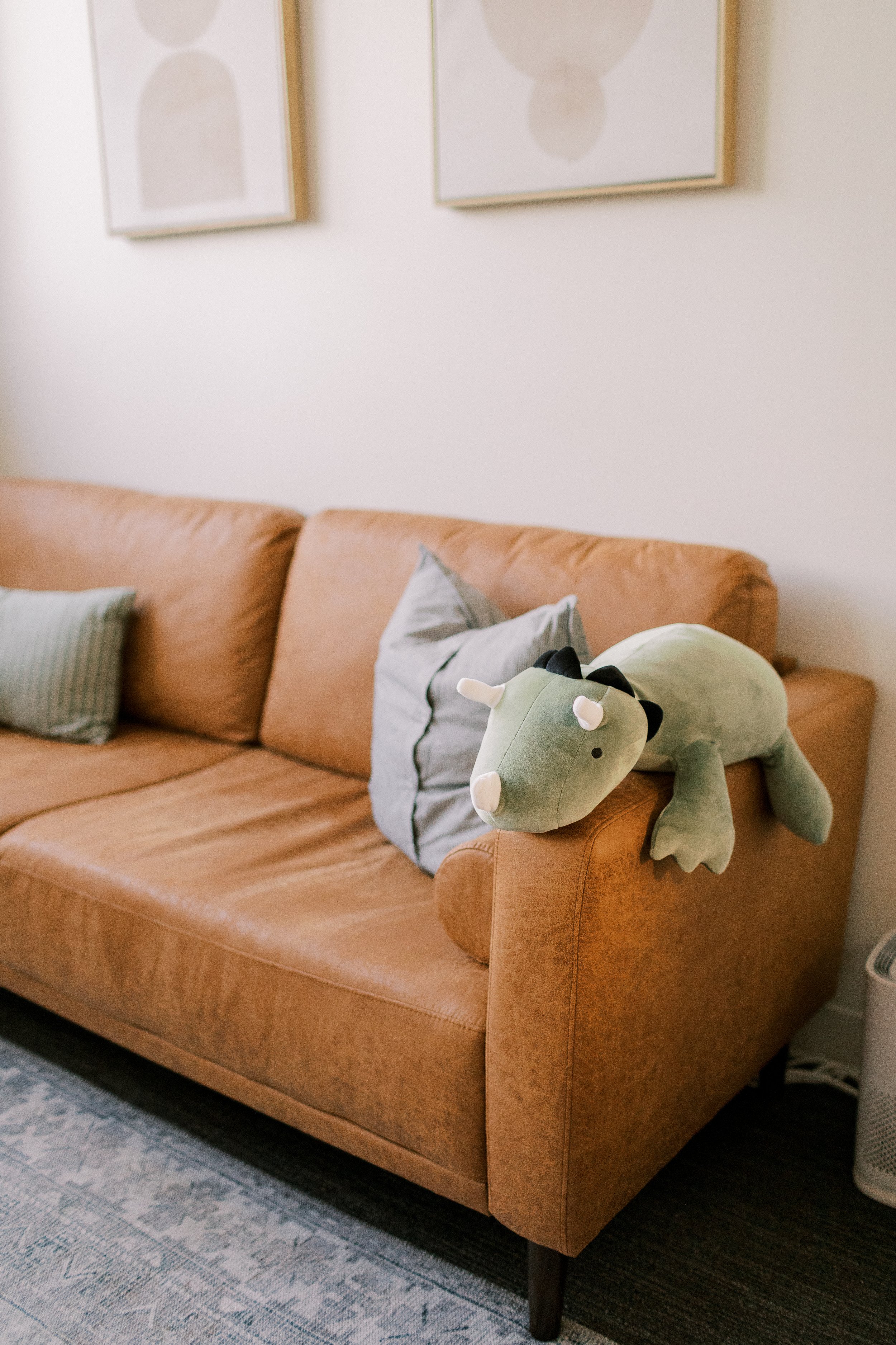 Living room with a tan sofa, decorative pillows, and a plush green dinosaur toy resting on the armrest. Wall art hangs above the sofa, and a patterned rug covers the floor.