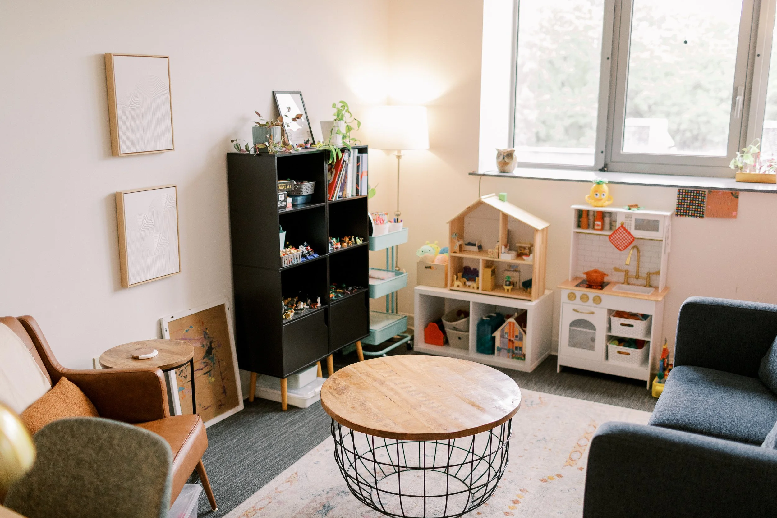 A cozy living room with a circular wooden coffee table, brown leather armchair, gray upholstered chair, black shelving unit filled with toys, toys on the floor, a small white play kitchen, and a window with a cat figurine on the windowsill. Light from the window illuminates the space.