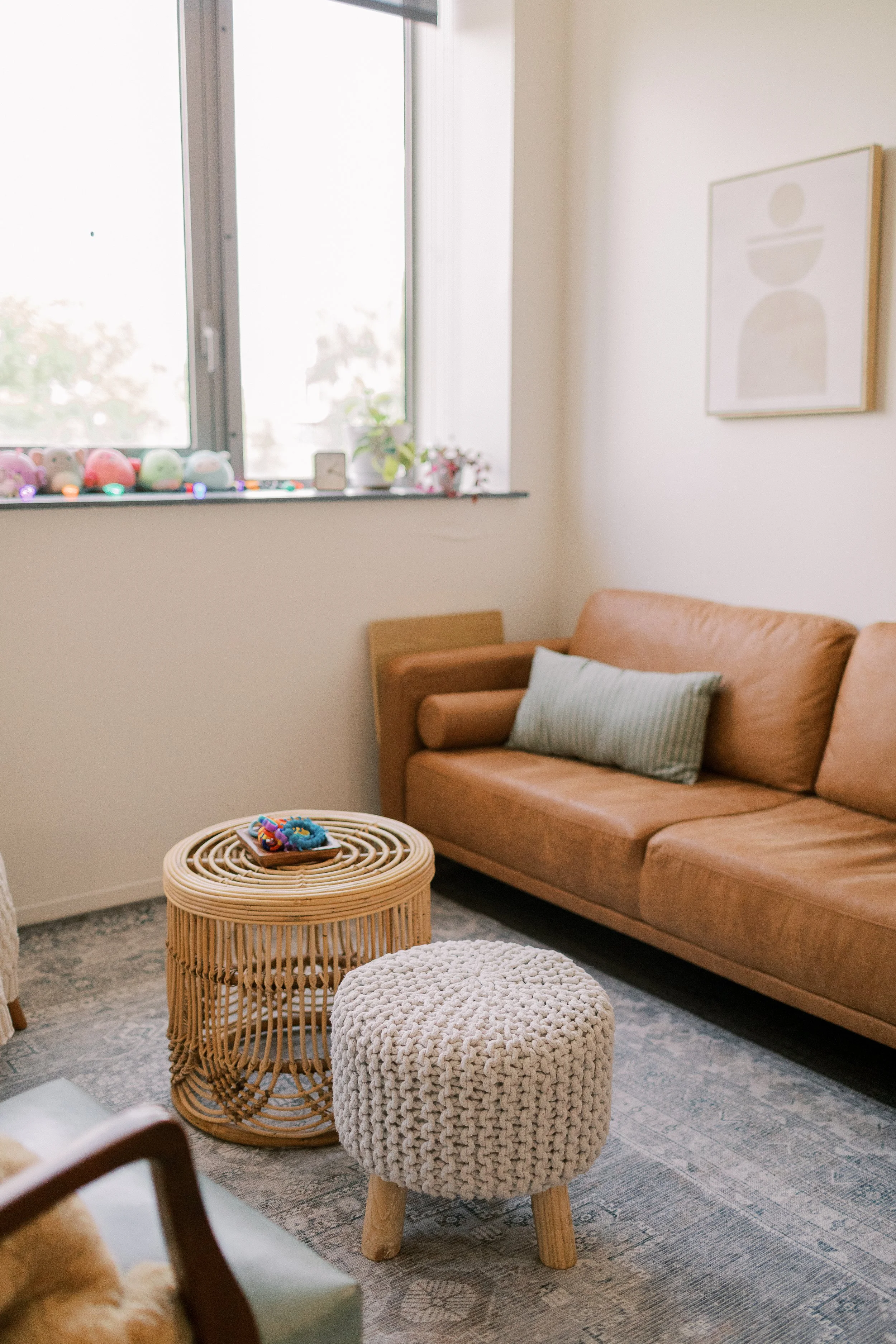A cozy living room corner with a tan sofa, a round wicker side table, a knitted pouf, a gray rug, and a artwork on the wall. A window with decorations and plants is in the background.