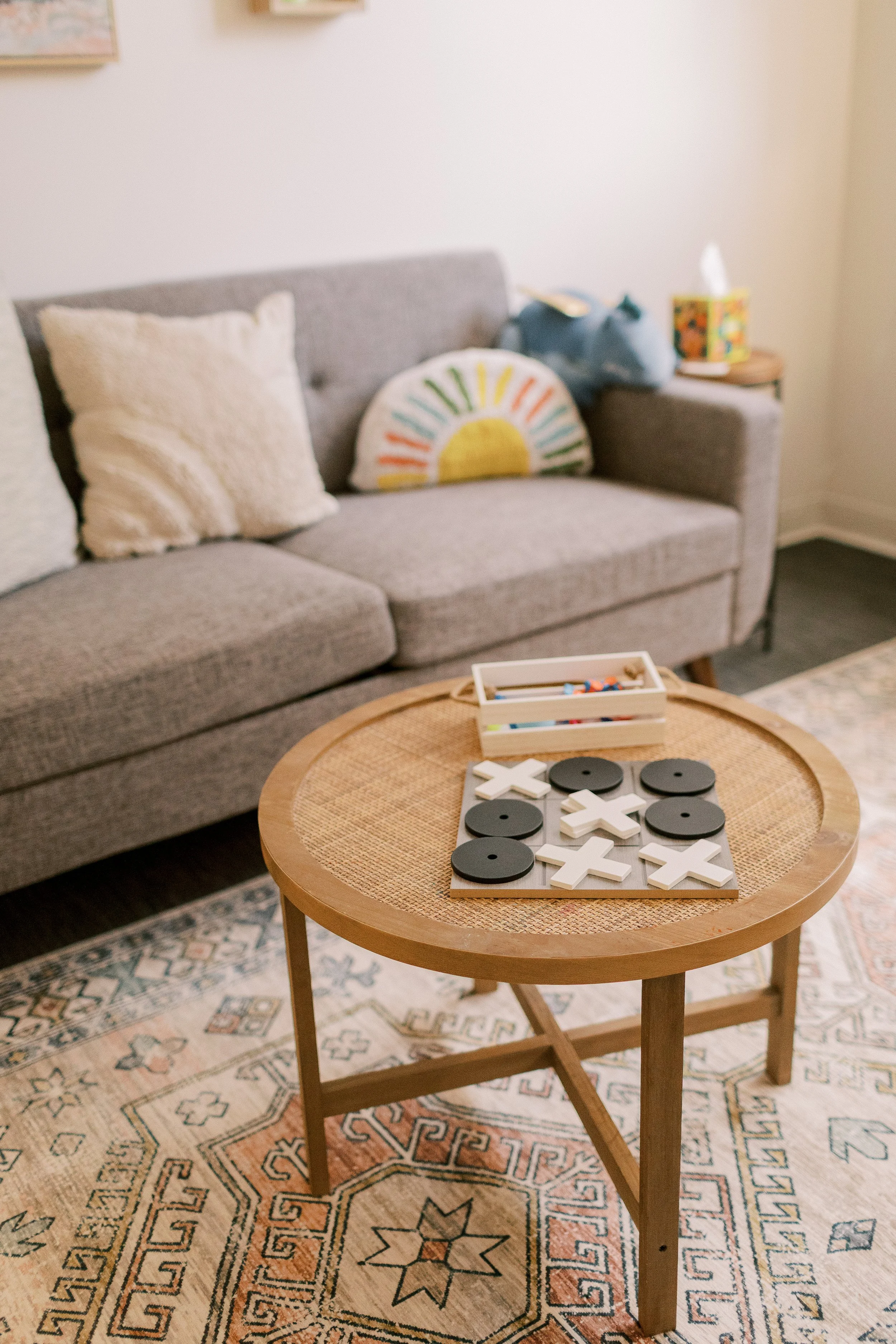 Living room with a gray couch, beige pillows, a colorful half-circle pillow, a blue plush elephant, and a nightstand with a tissue box. A wooden tray table with tic-tac-toe game pieces on a patterned rug.