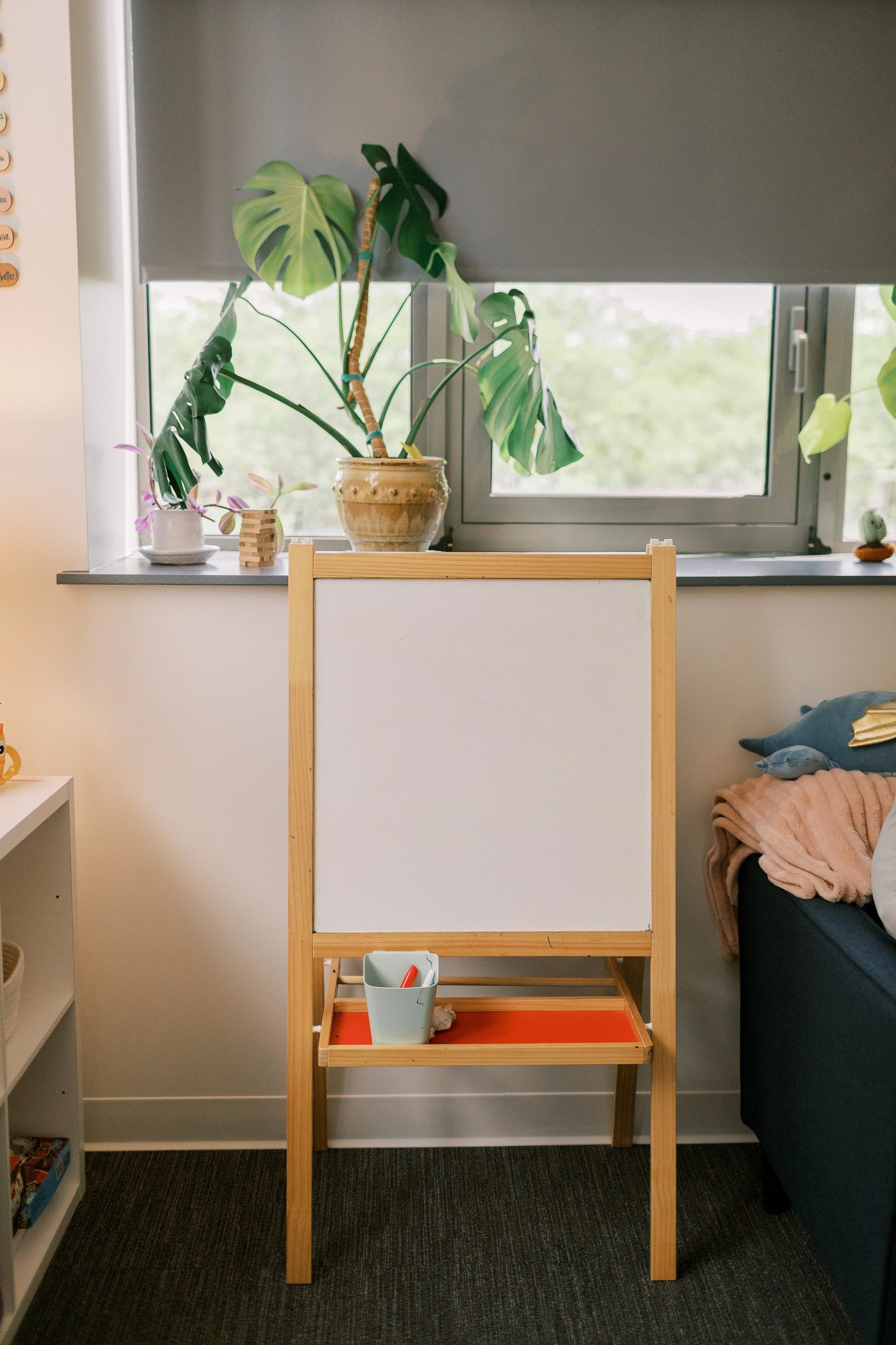 A wooden easel with a whiteboard, standing in a room with a dark carpet and a window behind it showing green trees outside.