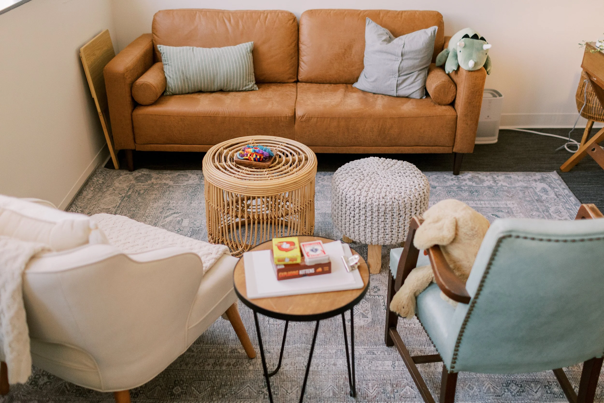 Living room with a tan leather couch, decorative pillows, a rattan side table, and a small round knitted ottoman in front. Various objects including books, a toy, and a plush toy are on the furniture and floor.