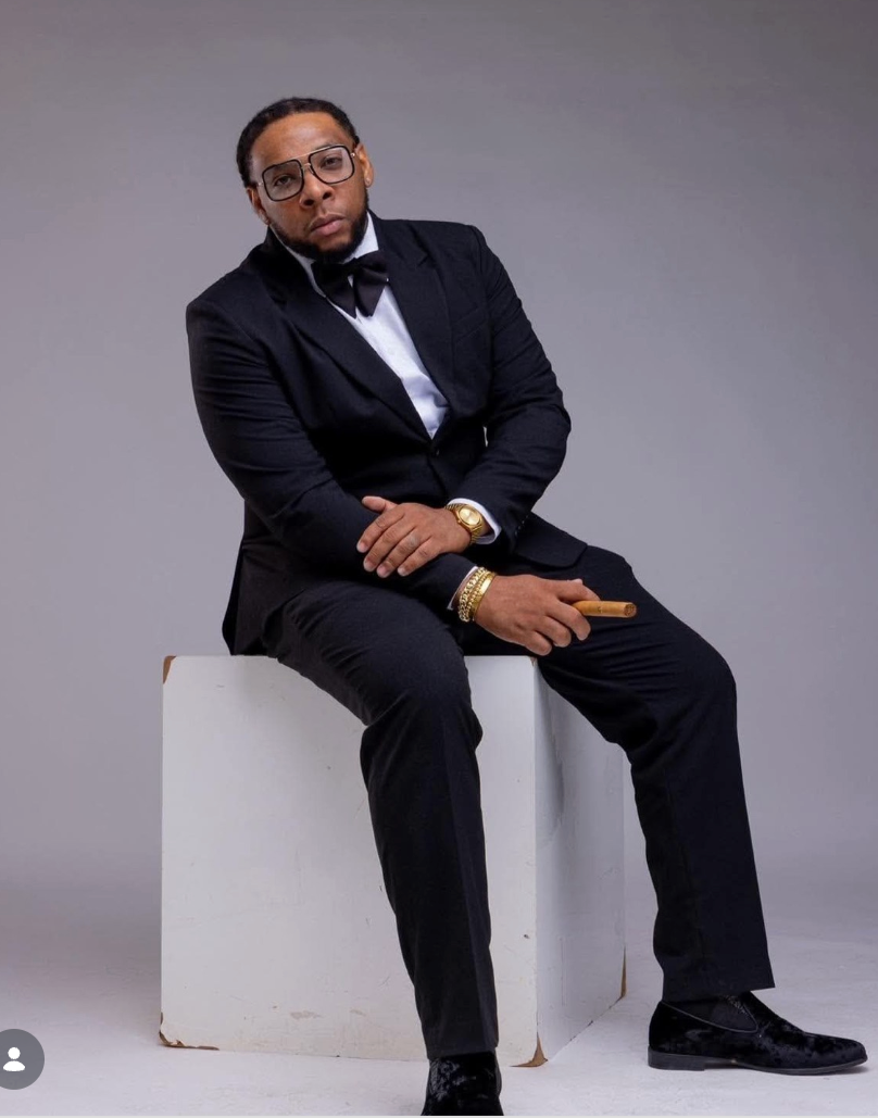 Tricky Hudson in a black tuxedo and glasses sitting on a white cube while holding a cigar