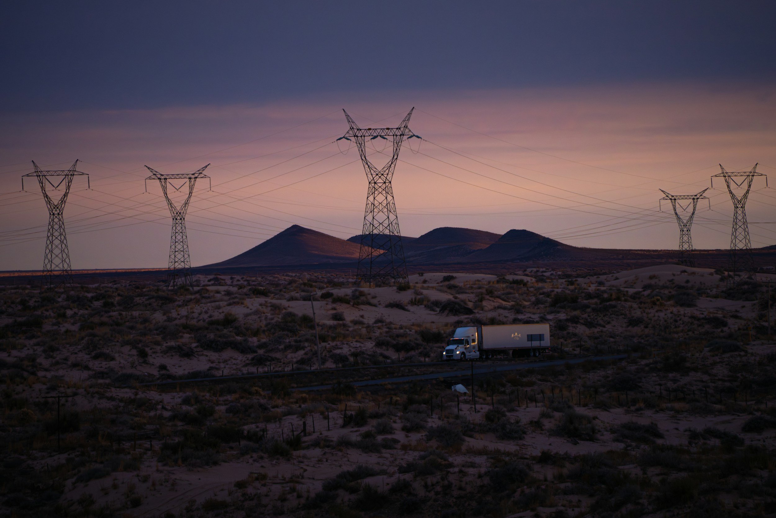 Sunset over a desert landscape with rolling hills, power lines, and a white semi-truck traveling on a highway.