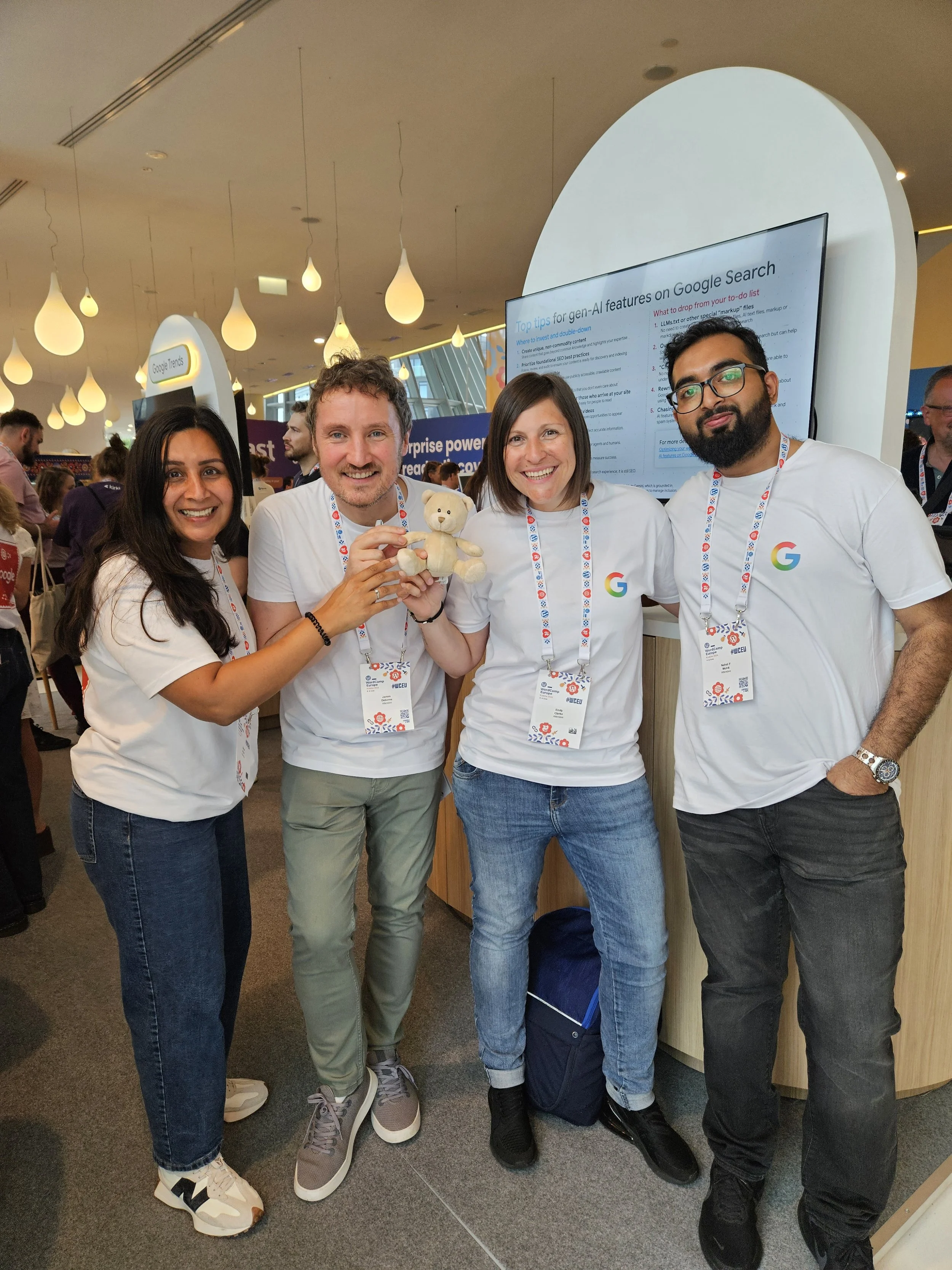 Four people smiling at a conference, holding a stuffed bear, wearing conference badges, standing in front of a display screen with presentation slides.
