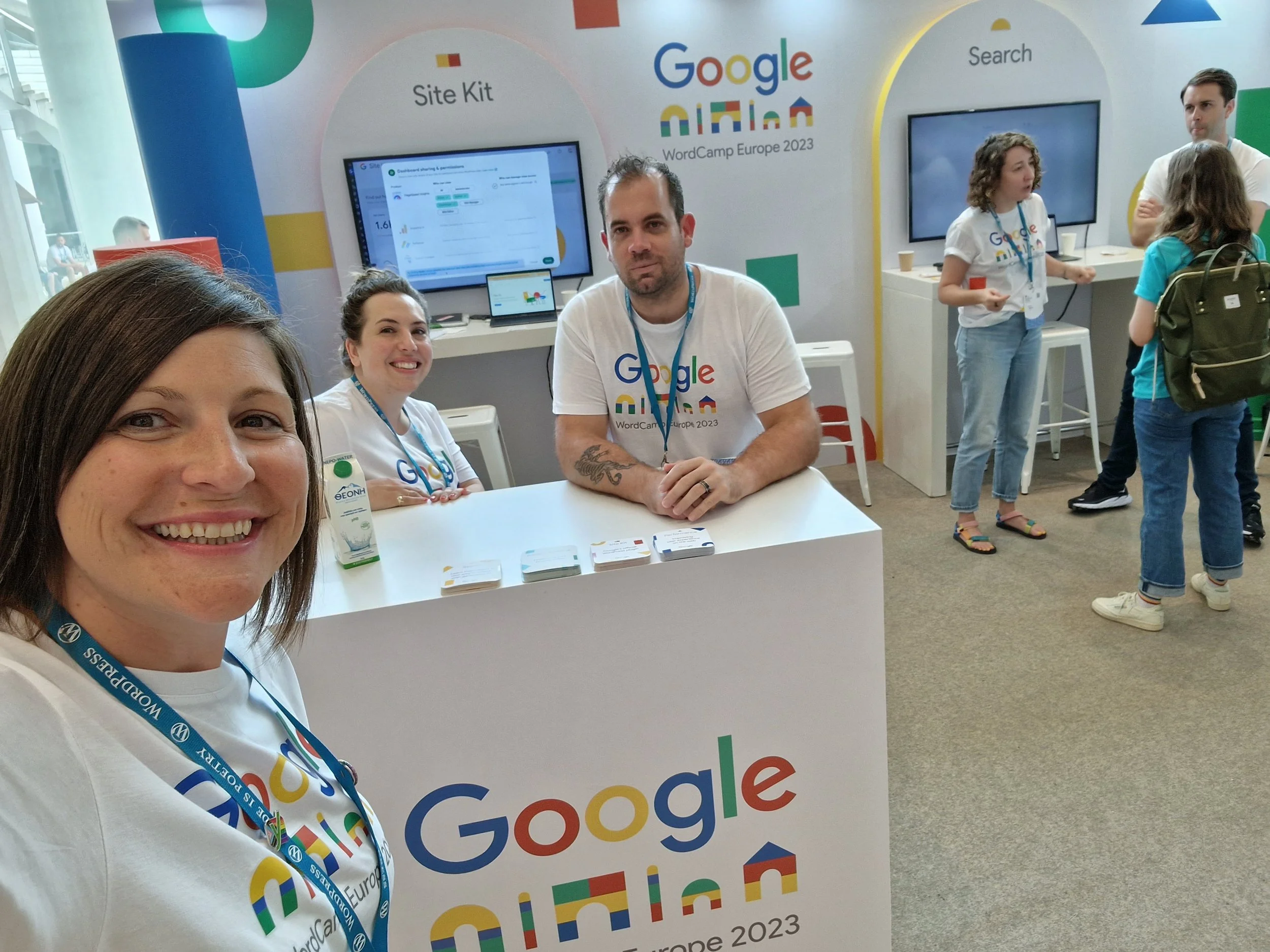 Group of people at Google WordCamp Europe 2023 booth, with a woman taking a selfie in the foreground, two women and two men sitting and standing near display screens, showing materials related to Google and WordPress.