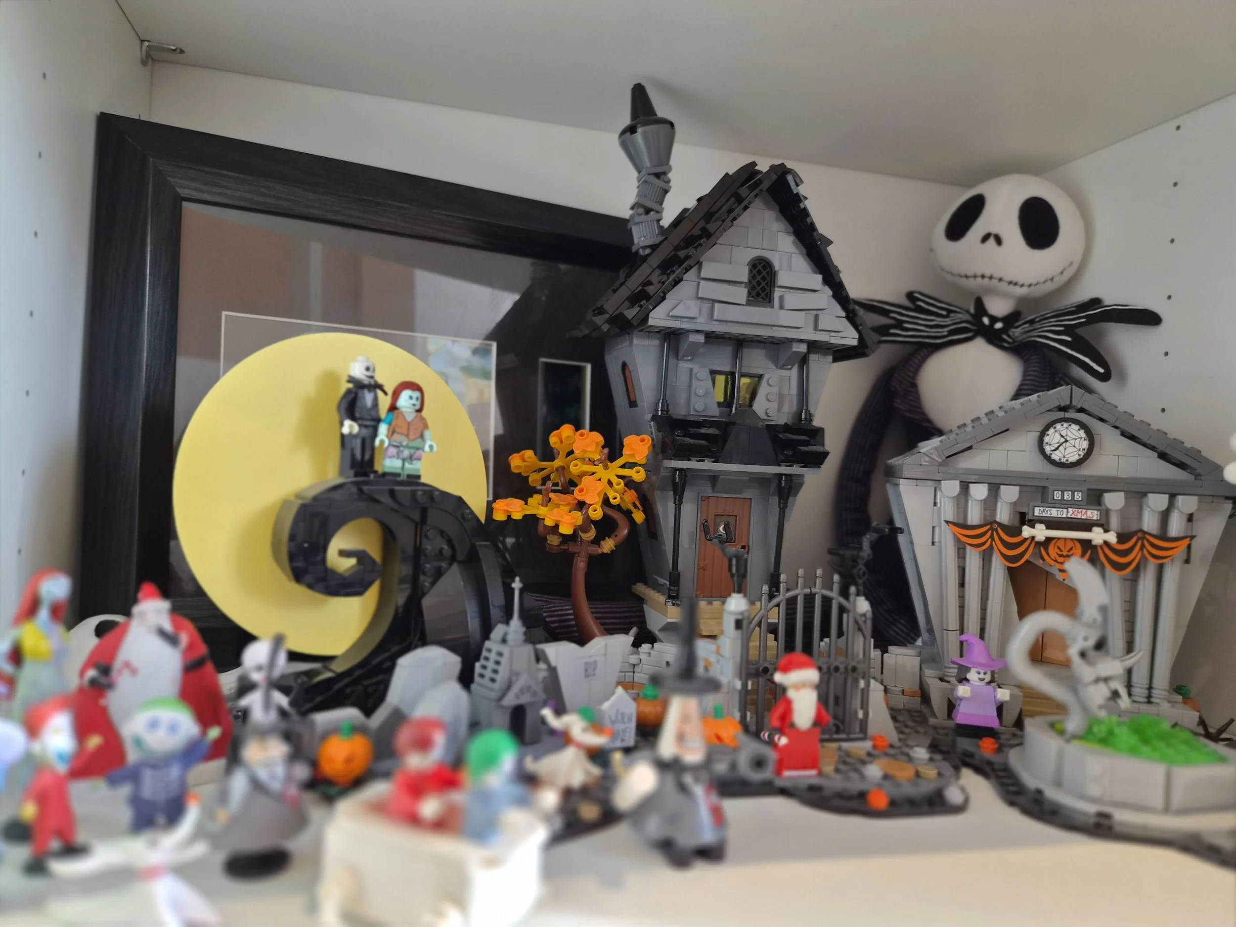 Collection of Halloween-themed LEGO decorations, including a haunted house, skeleton figure, scarecrow, pumpkins, ghosts, and Halloween characters, displayed on a shelf.