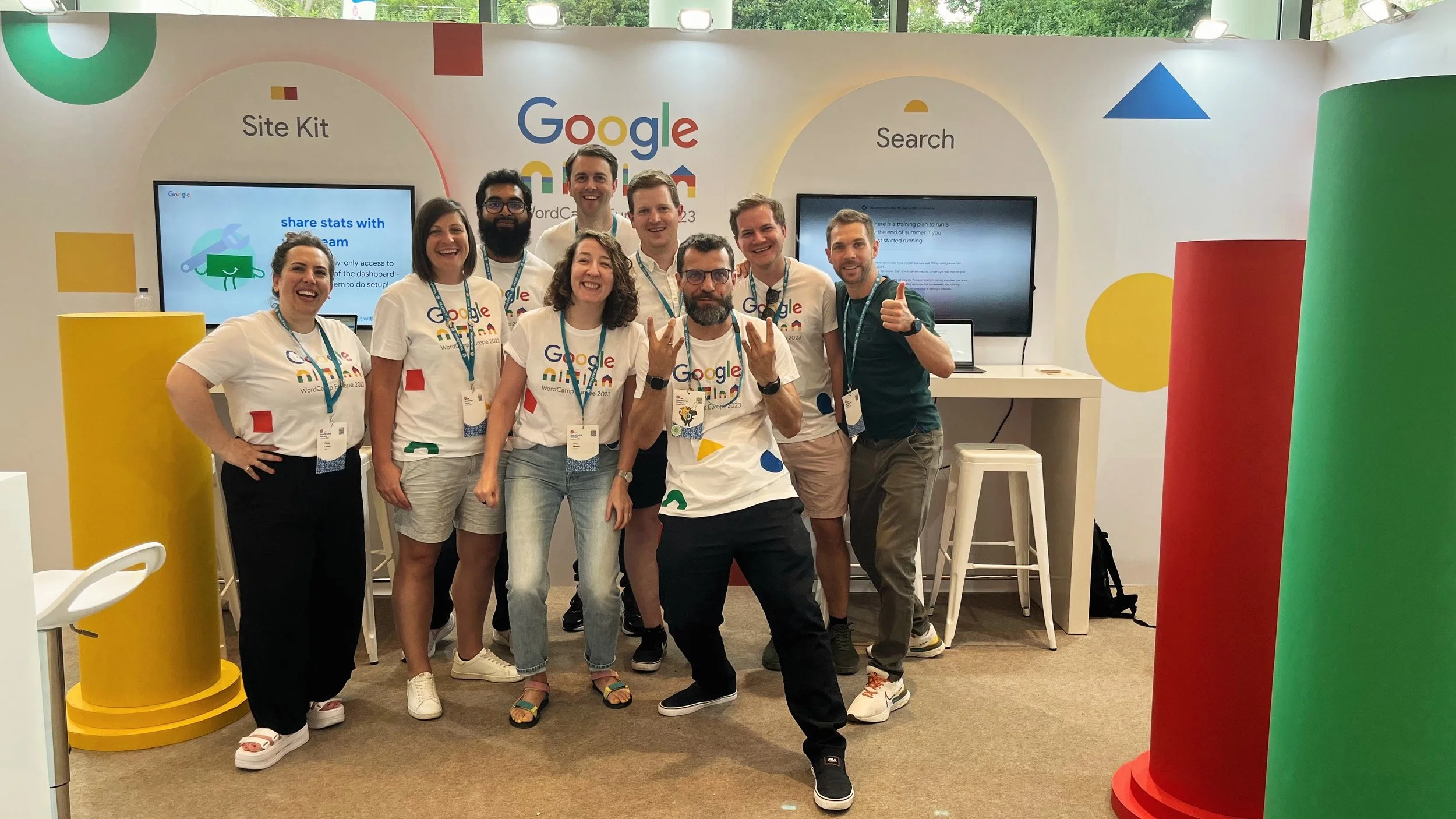 Group of people smiling and posing for a photo at a Google event, with a background displaying Google branding and large colorful cylindrical decorations.