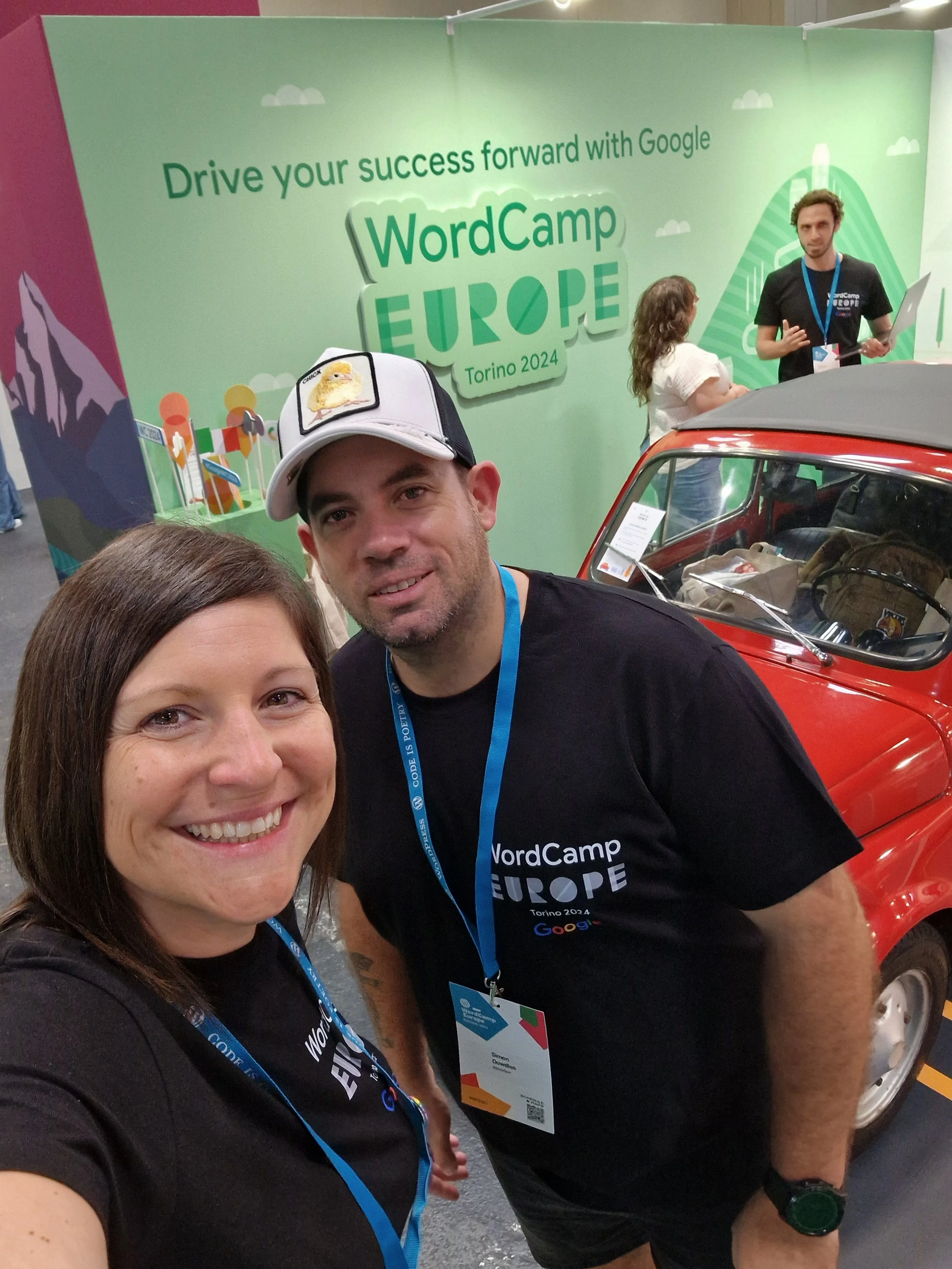 Two smiling people taking a selfie in front of a large green WordCamp Europe 2024 sign at a conference. Another person and a red vintage car are visible in the background.