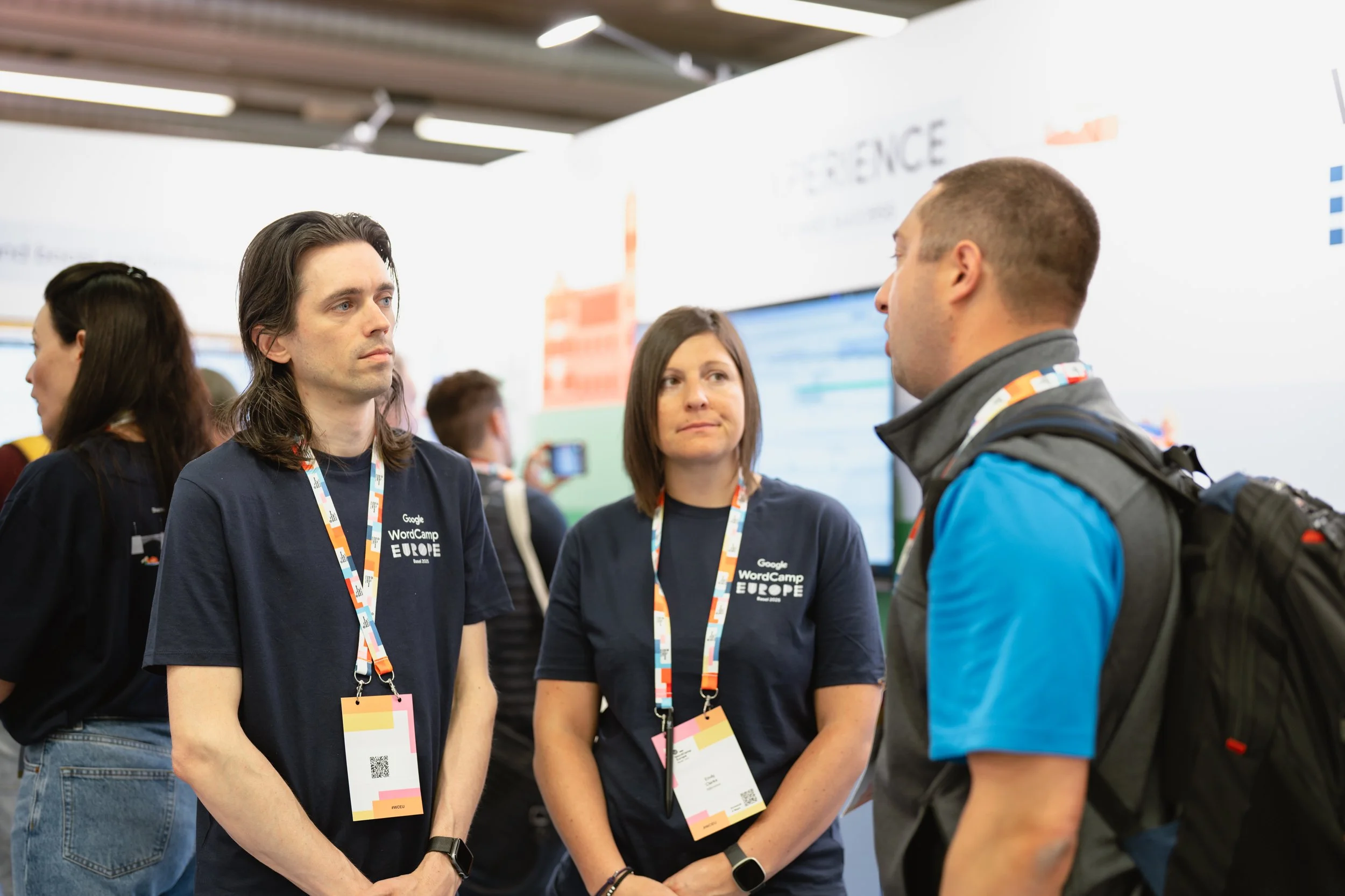 At a professional event, two women with lanyards and badges are engaged in conversation with a man. The women are wearing dark T-shirts with the text 'Google WordCamp Europe.' The background shows a booth or display area with banners and other attend
