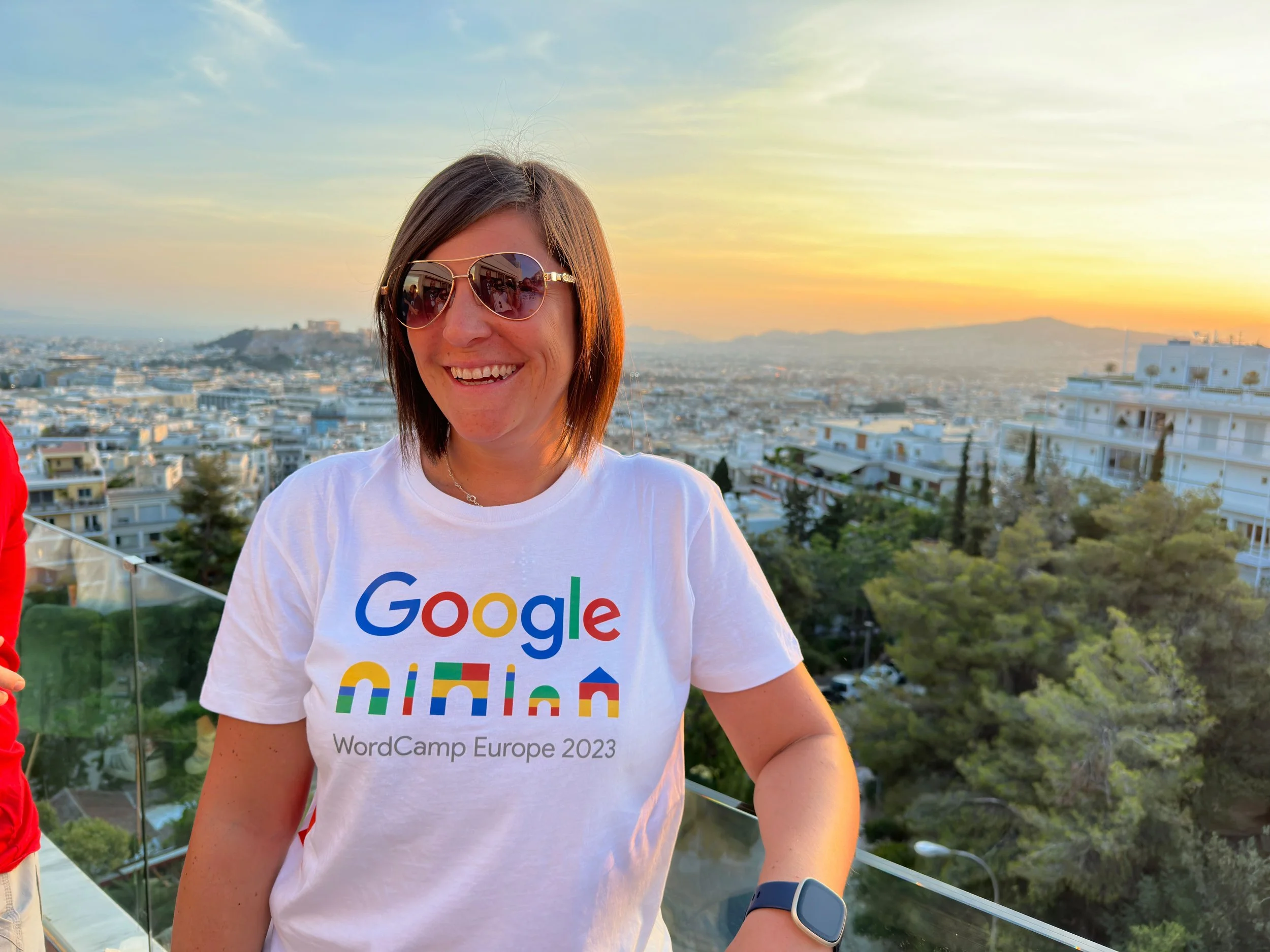 A woman wearing sunglasses and a Google-themed T-shirt at sunset, standing outdoors with a cityscape and hills in the background.