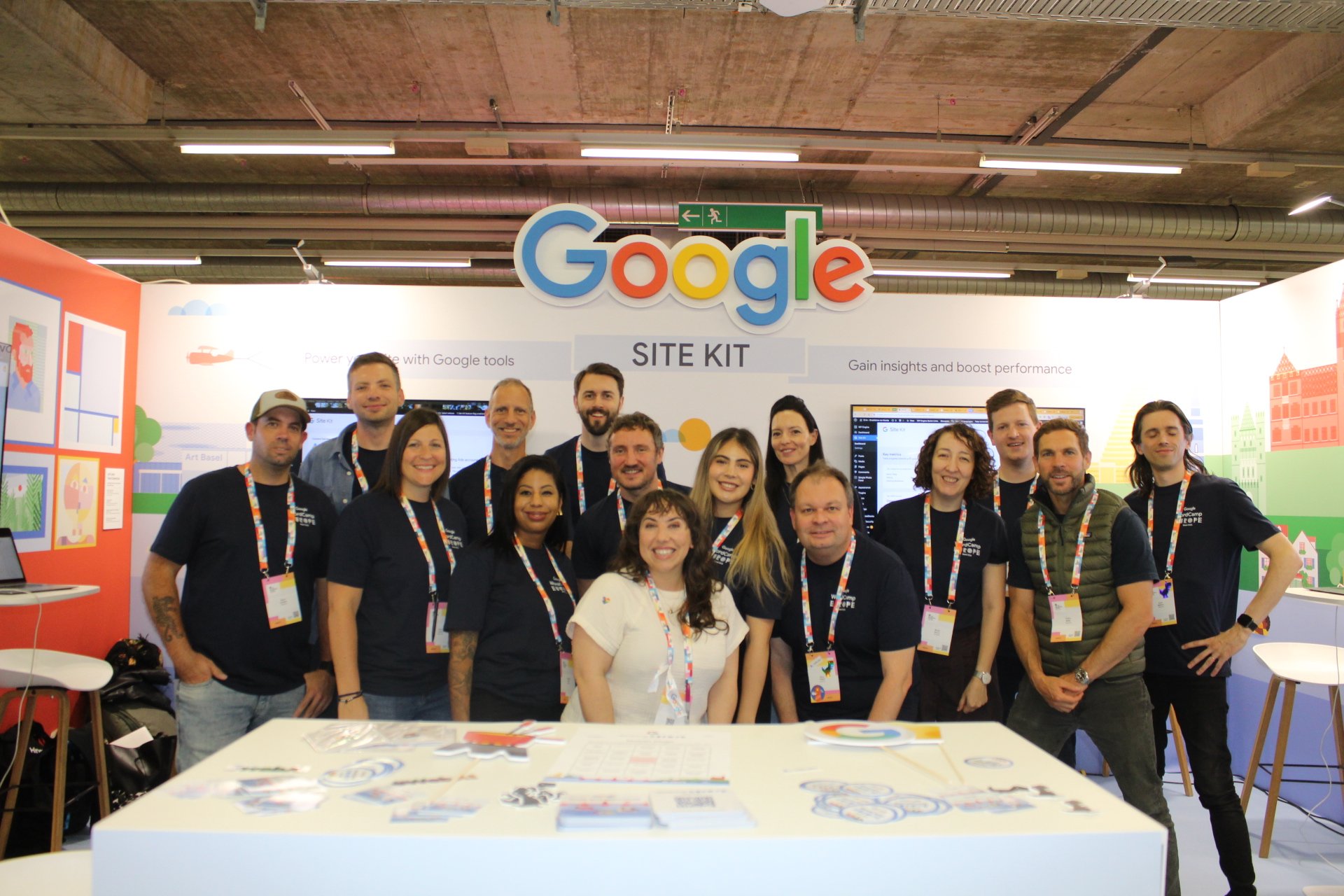 Group of people at a Google event, standing in front of a Google sign, smiling for the camera, with a display screen and colorful artwork in the background.
