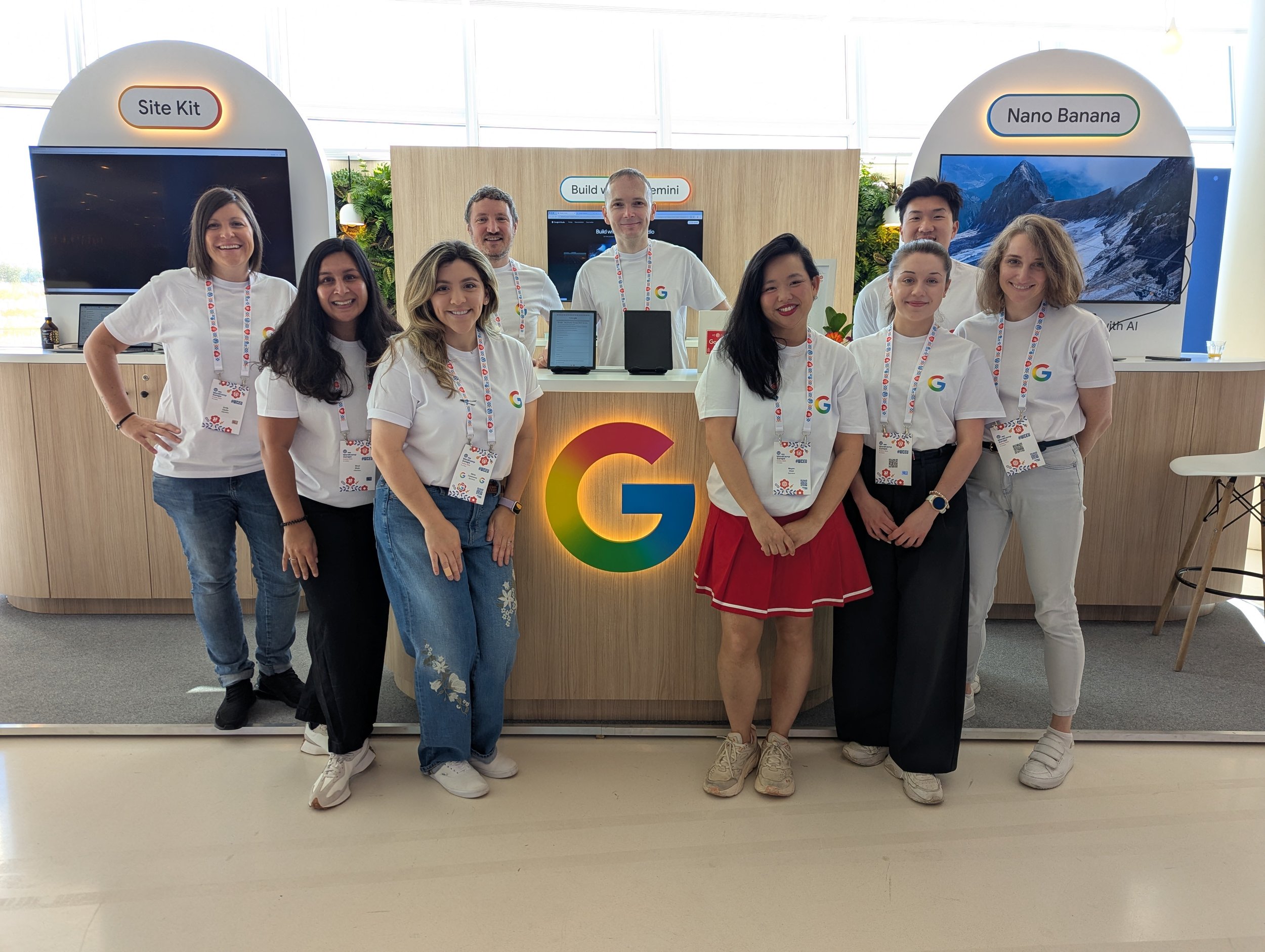 A group of nine diverse people, all wearing white Google's T-shirts and lanyards, standing indoors in front of a display with signs labeled "Site Kit," "Build with Gemini," and "Nano Banana." The setting appears to be a tech or corporate event.