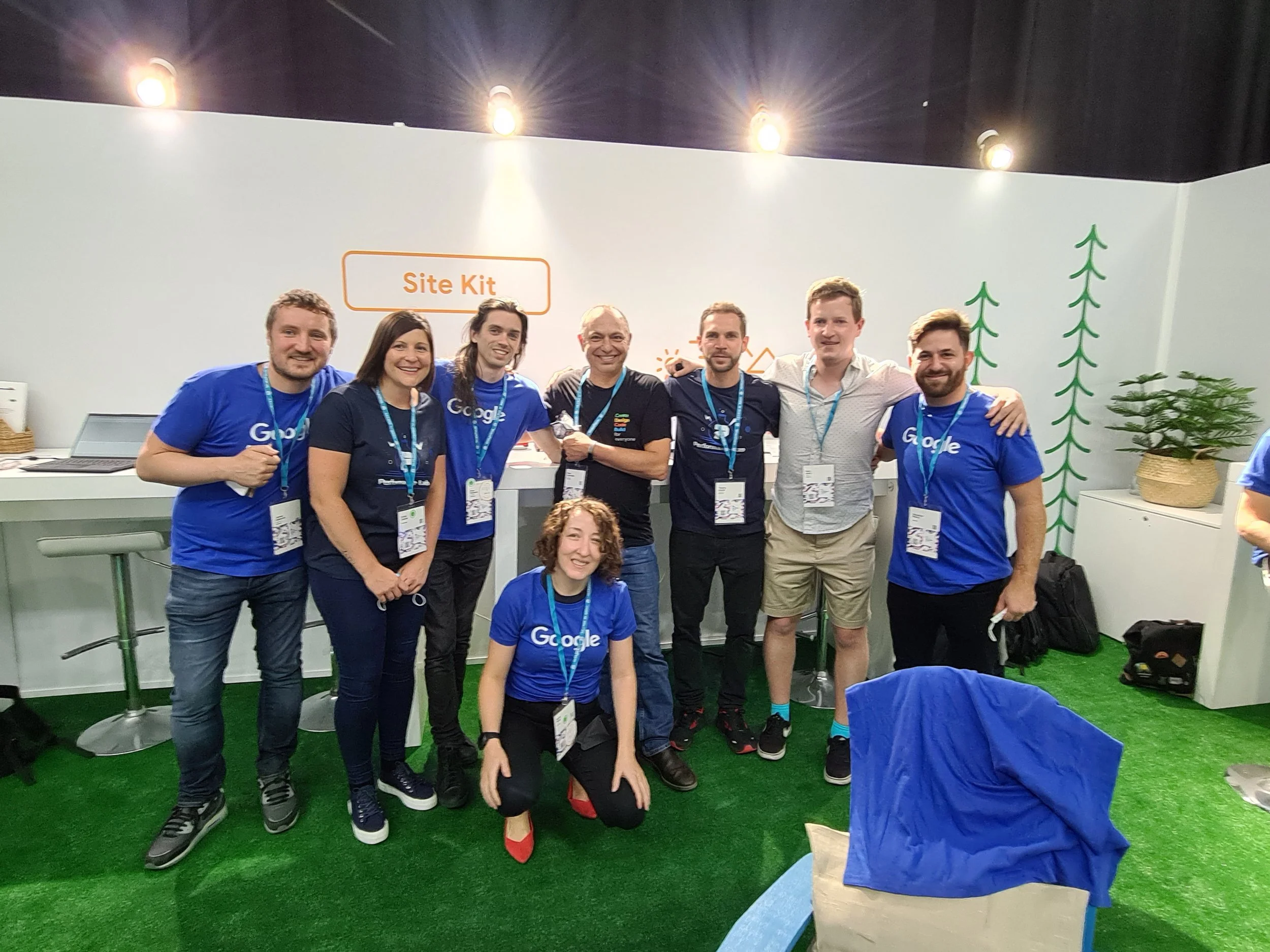 Group of people standing together at a Google event, smiling, with a white wall background and a sign that reads 'Site Kit'. Some are wearing Google-branded blue shirts, and there are potted plants beside them.