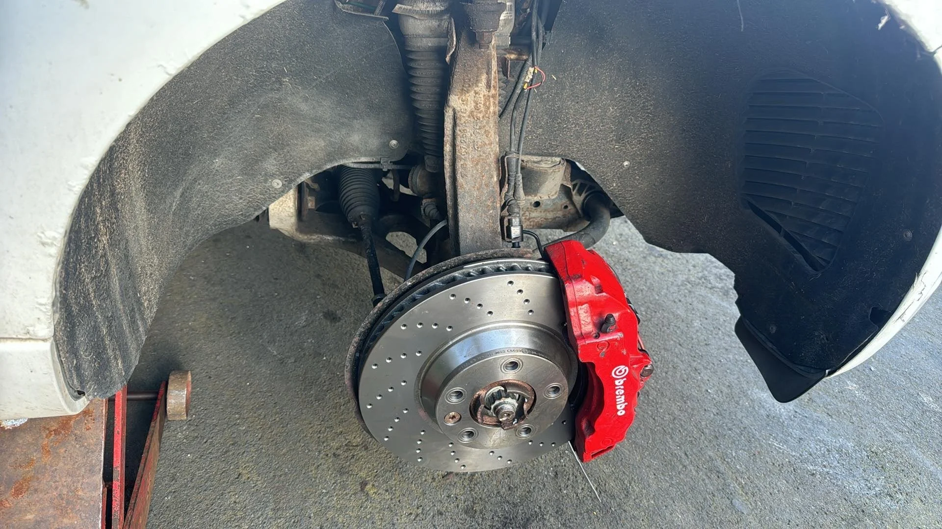 A car with the front wheel removed, showing a silver drilled brake rotor and a red Brembo brake caliper, on a vehicle under repair. The surrounding wheel well is dusty and the car is elevated on supports.