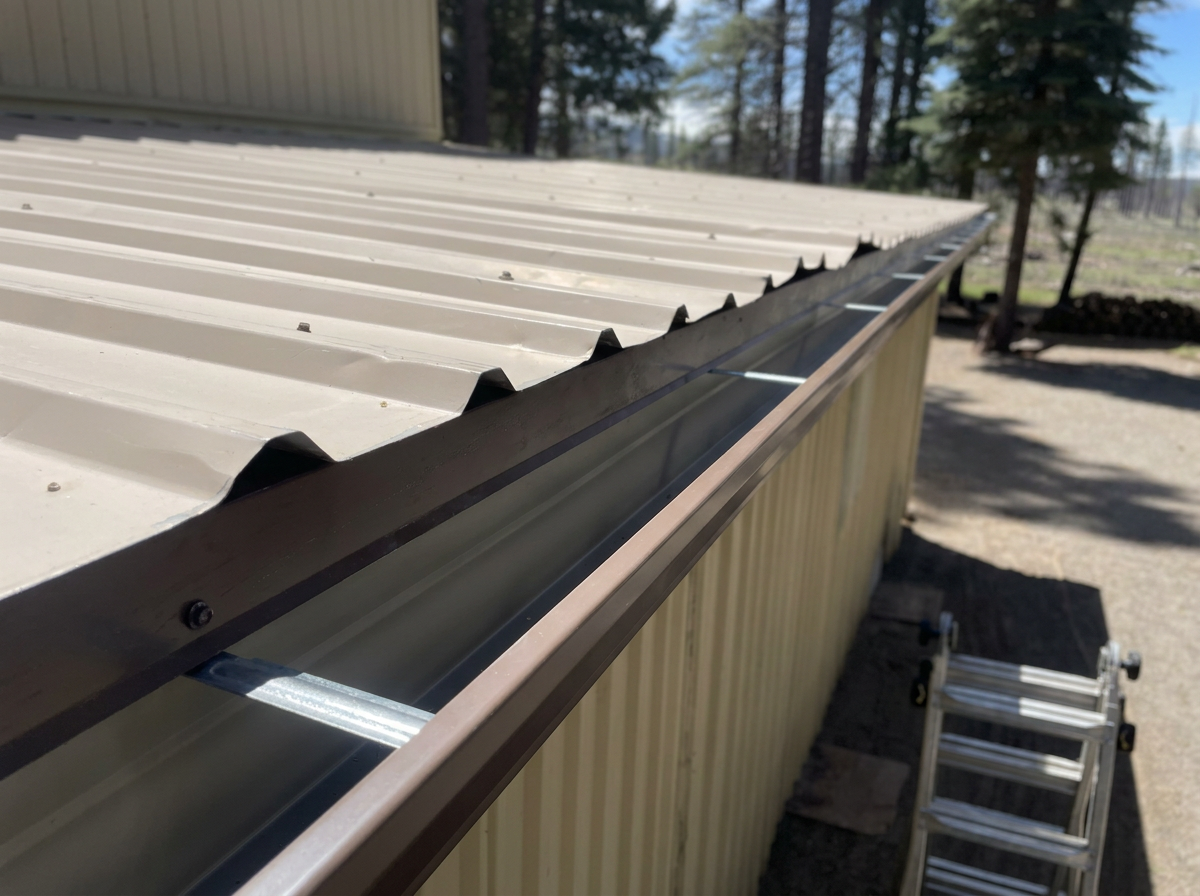 Close-up view of metal roofing panels with a gutter system along the edge of a building, with trees in the background.