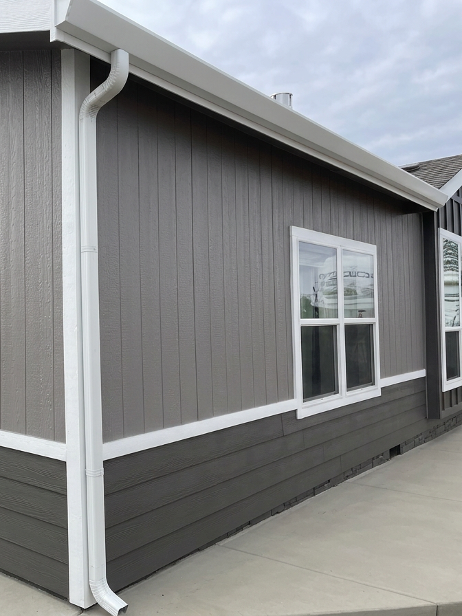 Close-up of a modern gray house exterior with a white gutter system and a double-pane window, on a cloudy day.