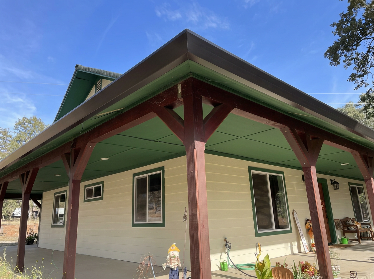 Front of a house with a porch, cream-colored siding with green trim around the windows and door, and a green roof. There are some outdoor decorations, potted plants, and furniture on the porch.