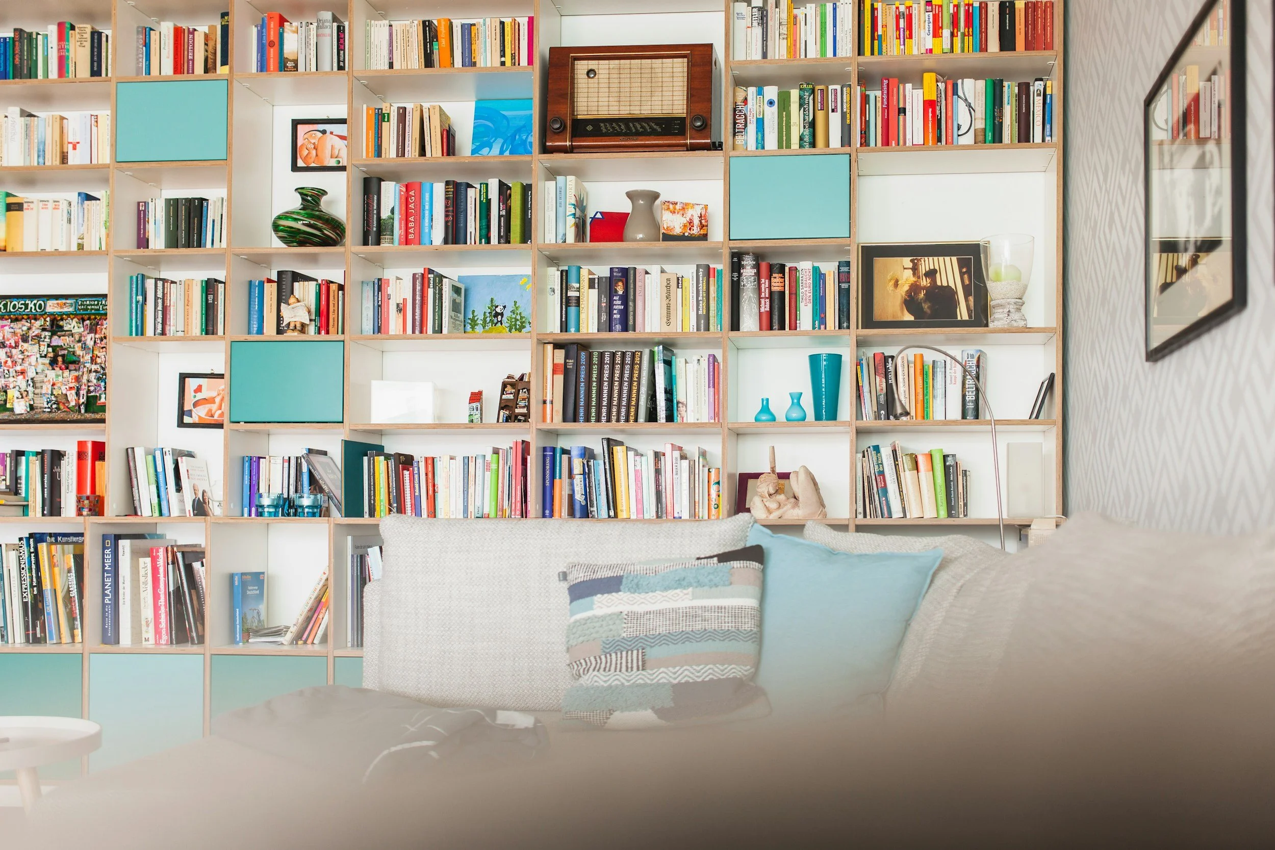 Close-up of a white sofa with a patterned pillow in front of a large wooden bookshelf filled with books, home decor items, framed photos, and vintage radio, with a partial view of a wall with patterned wallpaper.