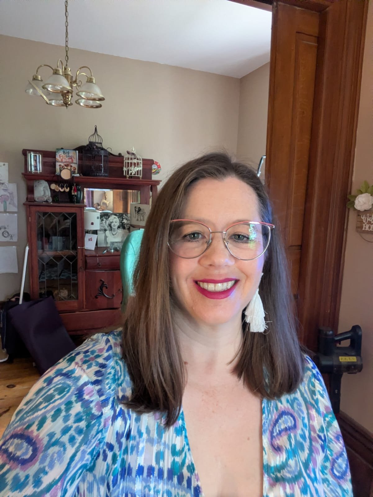 A woman with long brown hair, glasses, and earrings smiling in a room. Behind her is a wooden cabinet with a mirror, birdcages, and family photos. The room has beige walls, a chandelier, and wooden trim.