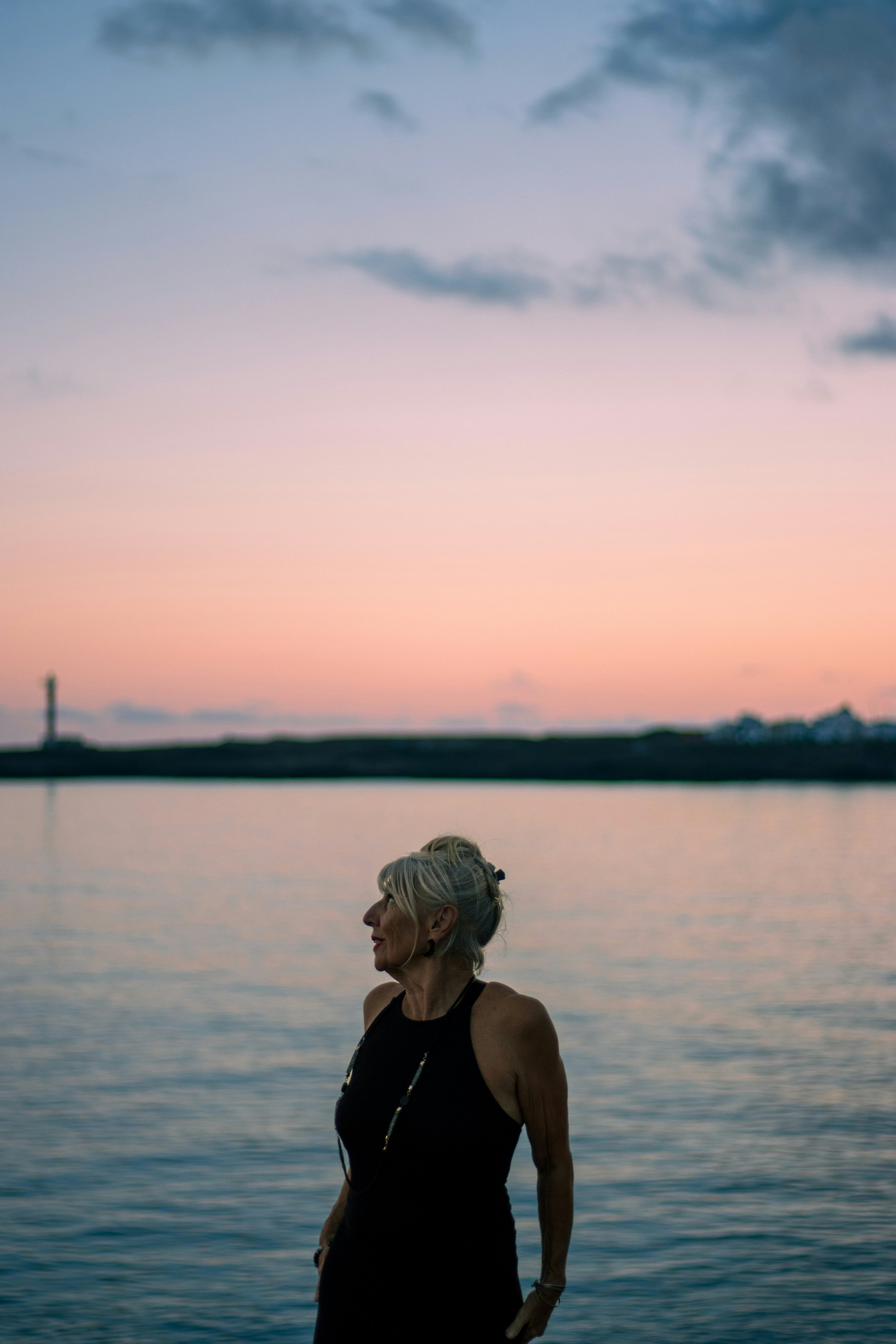 An older woman with gray hair stands by the water during sunset, with a colorful sky in the background.