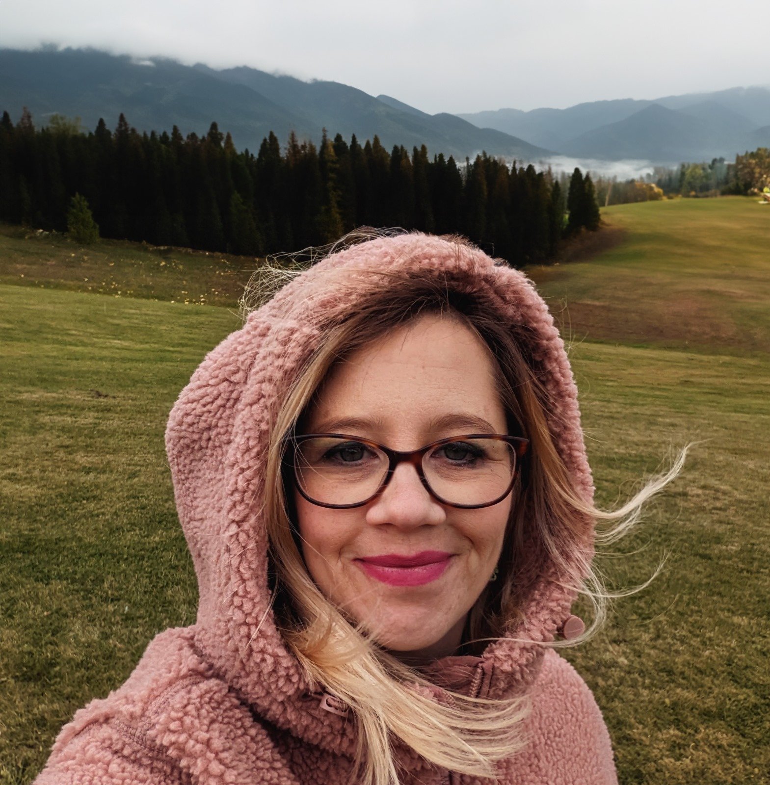 A woman wearing glasses and a pink fluffy hooded coat outdoors, with a backdrop of rolling green fields, dense forest, and mountains under a cloudy sky.