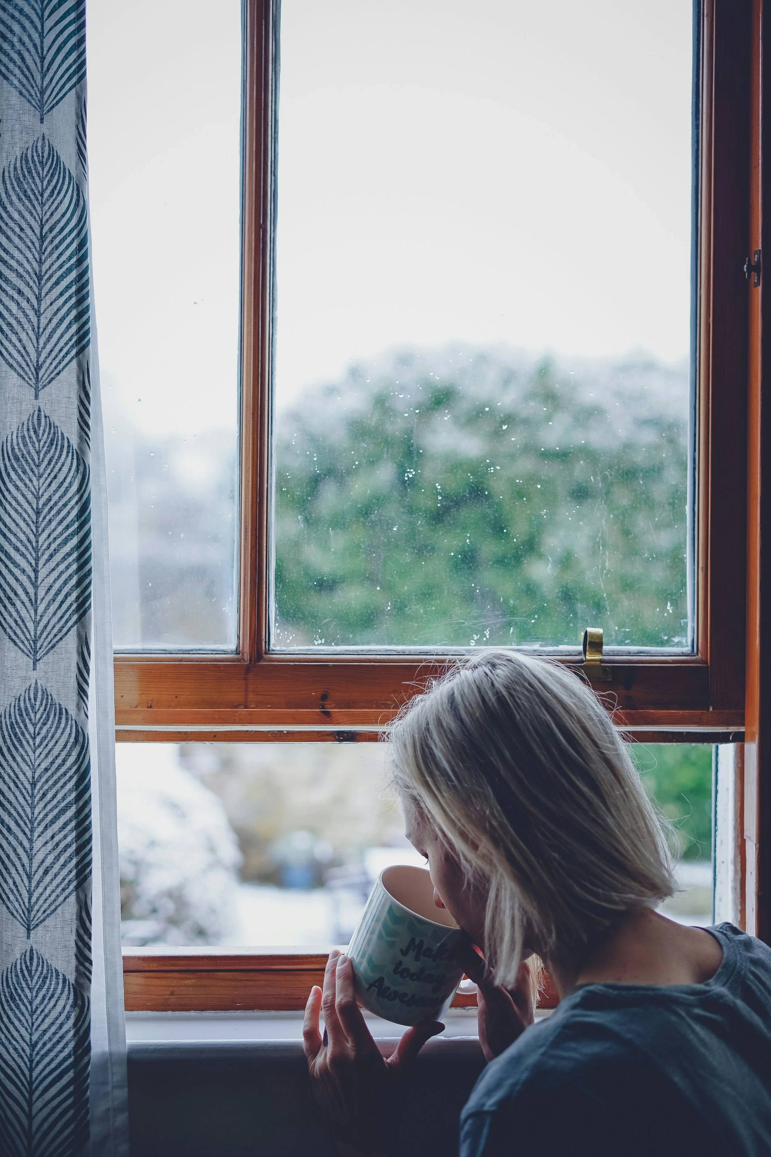A woman with blonde hair drinking coffee from a mug near a window with rain droplets on the glass, daytime, curtains on the side, trees blurred outside.