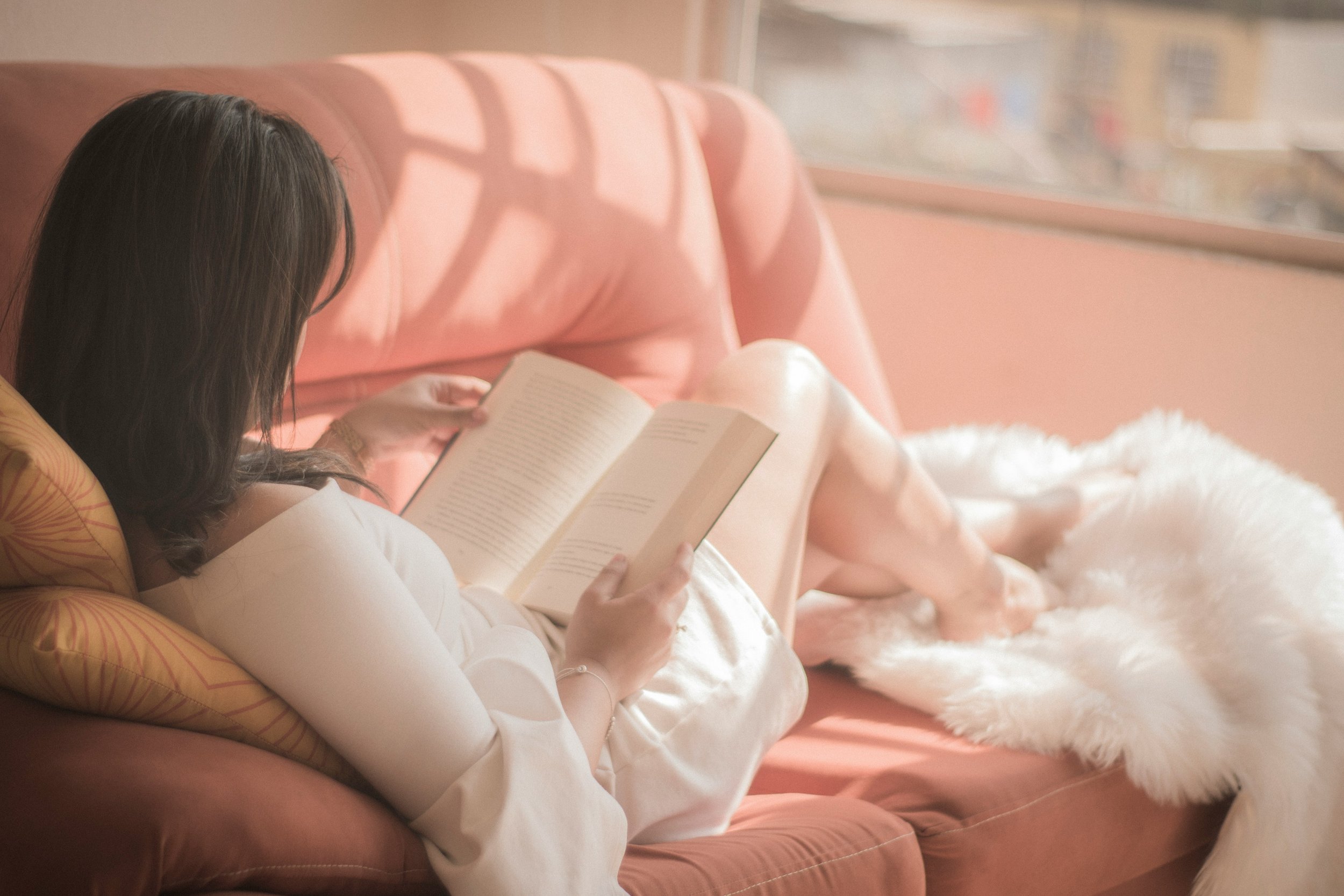 A woman with dark hair reading a book while sitting on a pink couch with orange and yellow patterned cushions. She is wearing a white long-sleeve top, and a white fluffy rug is on the floor next to her.
