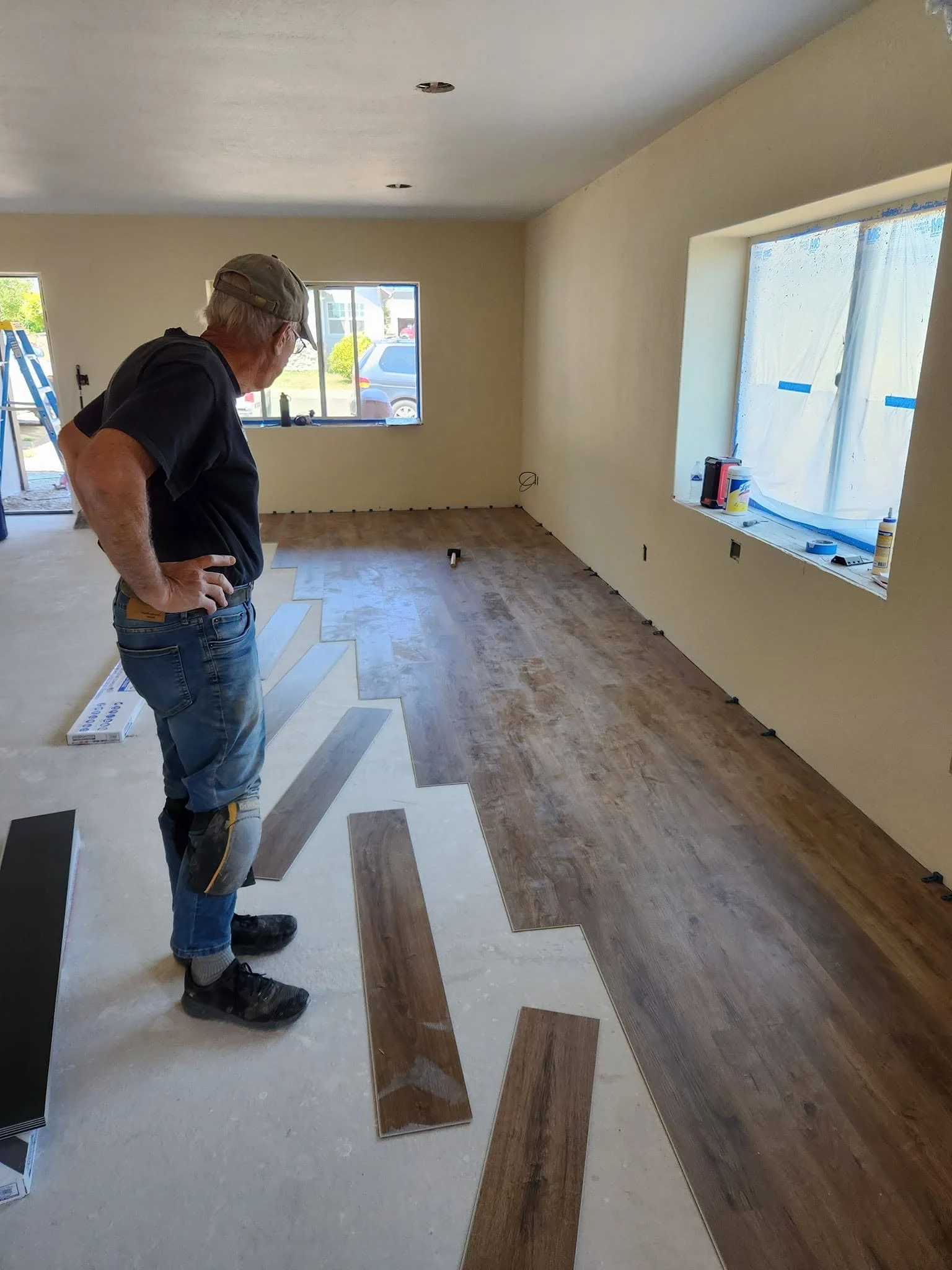 A man wearing a cap, black shirt, and jeans is installing or selecting flooring tiles inside a room with large windows, some of which are covered with plastic sheeting, in a house under renovation or construction.