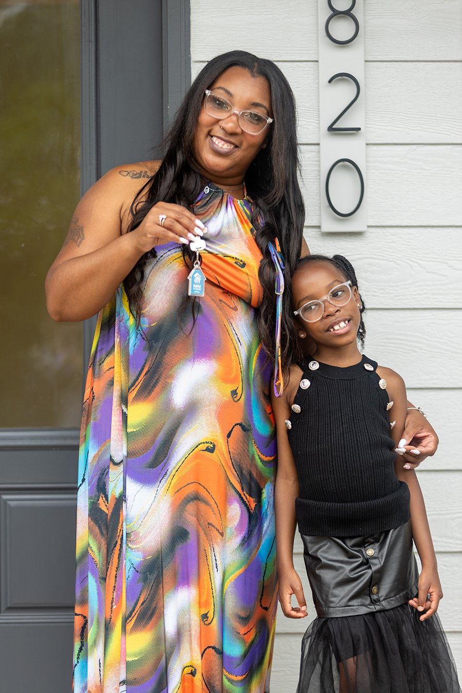 A woman and a girl standing outside a house. The woman is holding a key and smiling, while the girl is leaning against her. There is house number 820 on the wall behind them.