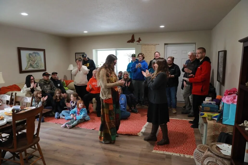 People gathered in a living room for a celebration, with some standing, some sitting on the floor, and children seated on the carpet. The room has artwork on the walls, and gifts and party supplies nearby.