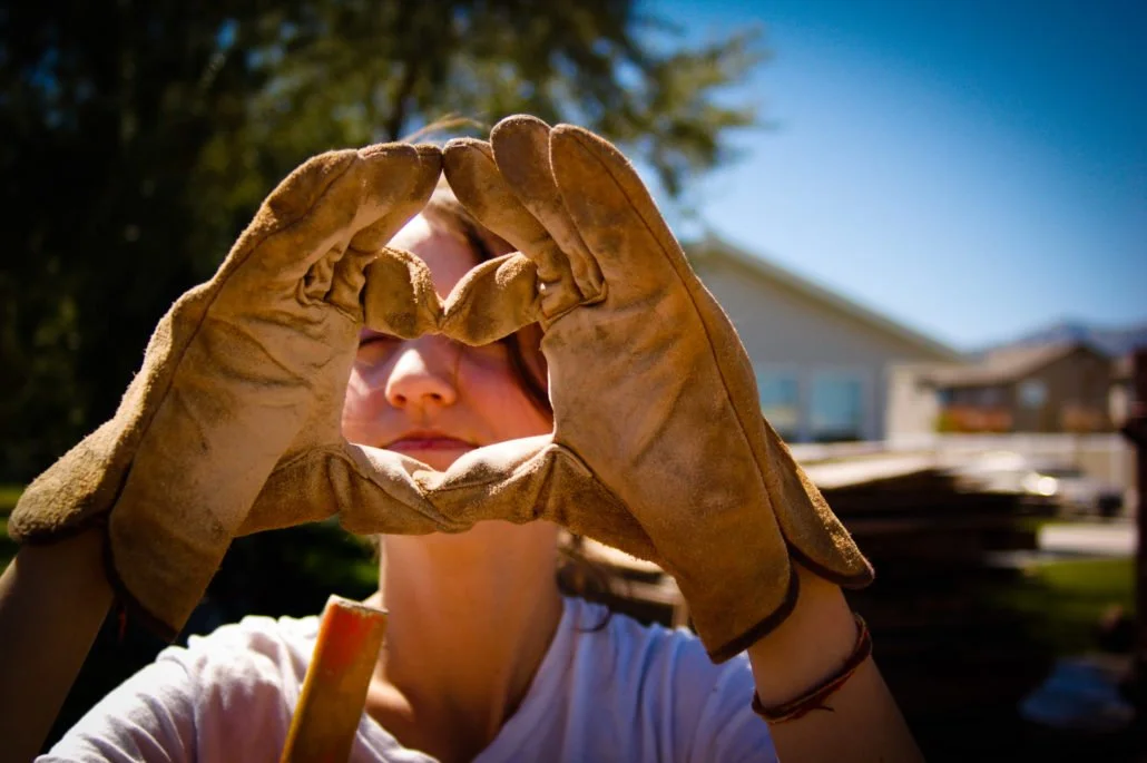 A person wearing work gloves making a heart shape with their hands outdoors, with a house and trees in the background.