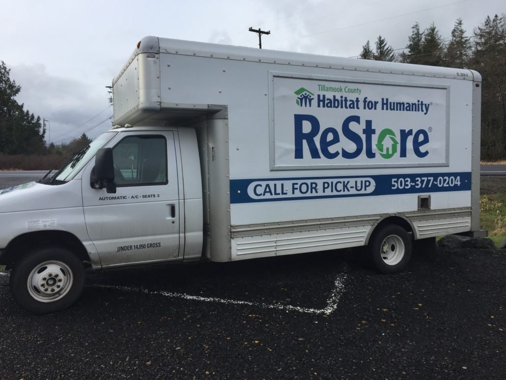 A white box truck with a Habitat for Humanity ReStore advertisement and contact information parked on a paved lot with a natural background.