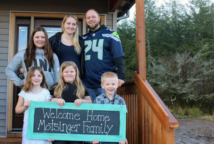 A family of seven standing on the porch of their home, holding a sign that says, 'Welcome Home Motsinger Family,' with trees and greenery in the background.