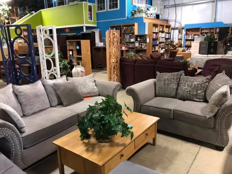 Furniture display in a store including two light grey sofas with pillows, a wooden coffee table with a potted plant, and a bookshelf background with various books and decorative items.