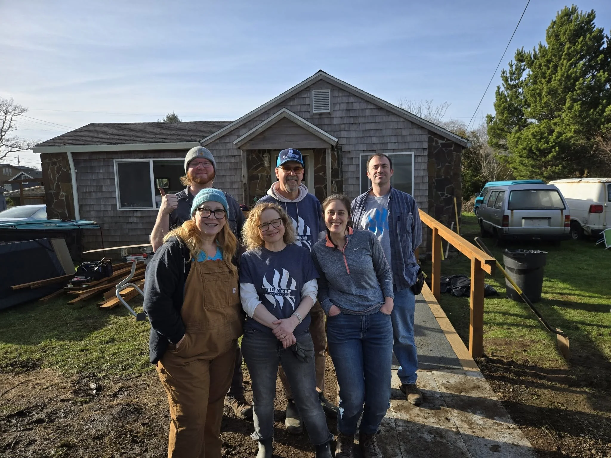 A group of seven people standing outside in front of a house, smiling and posing for the camera, with some construction or renovation work in progress on the house.