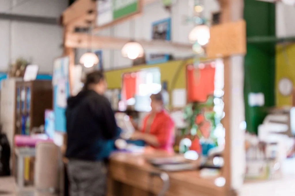Two people in a colorful indoor space, possibly a craft store or cafe, engaged in conversation, with various shelves and lighting fixtures in the background.