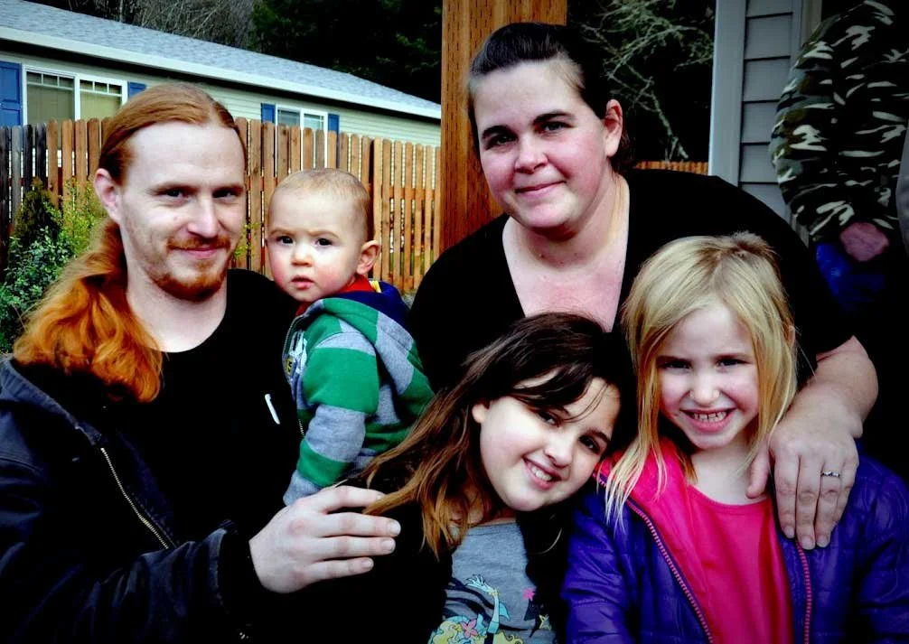 A family of five outdoors, with a man with long red hair, a woman, two young girls, and a toddler boy. They are smiling, standing in front of a wooden fence and house.