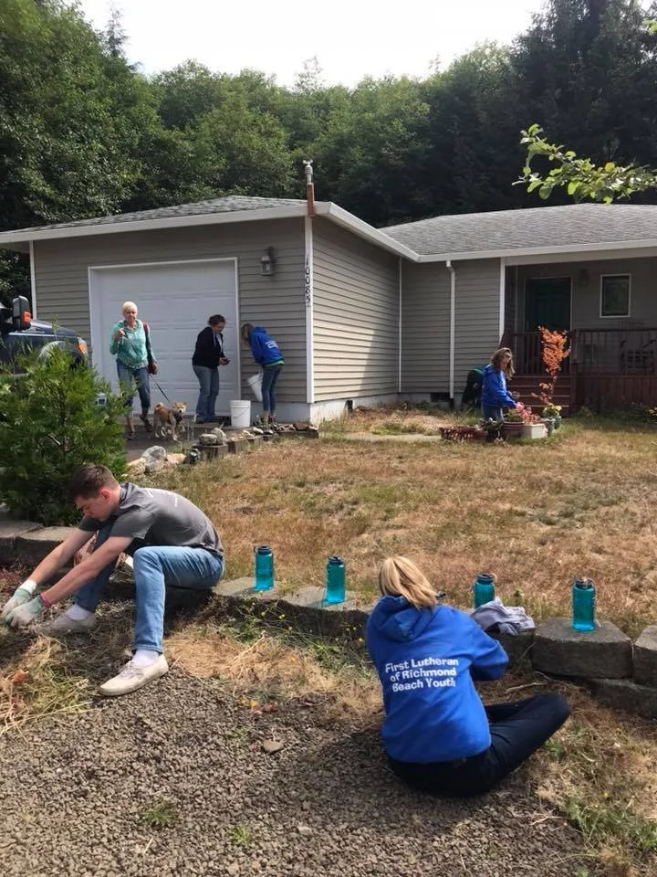 Group of people working on outdoor home projects in a yard with a house in the background.