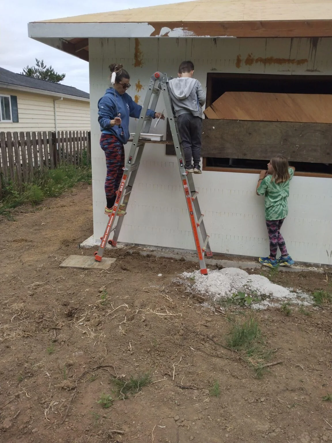 A woman and two children working on the exterior of a house, with the woman standing on a ladder and the children observing. The house's wall is partially covered with insulation or siding materials, and the ground is dirt with some scattered grass.