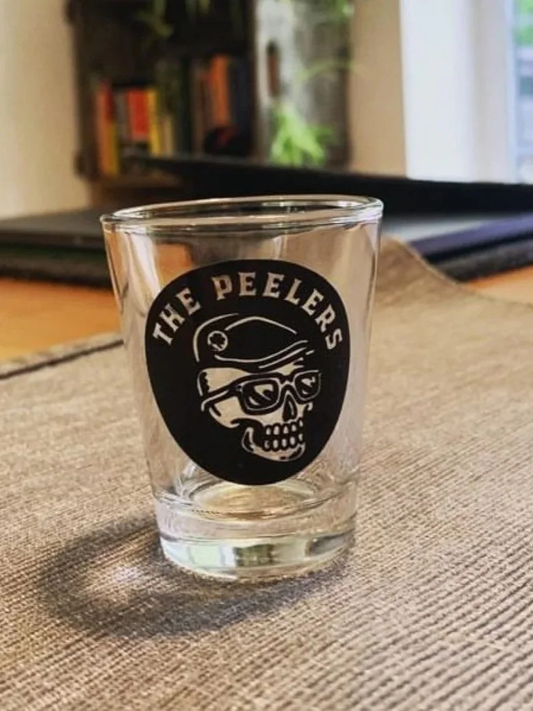 A clear glass with a skull wearing sunglasses and a helmet graphic, and the text 'THE PEELERS' on it, placed on a table.