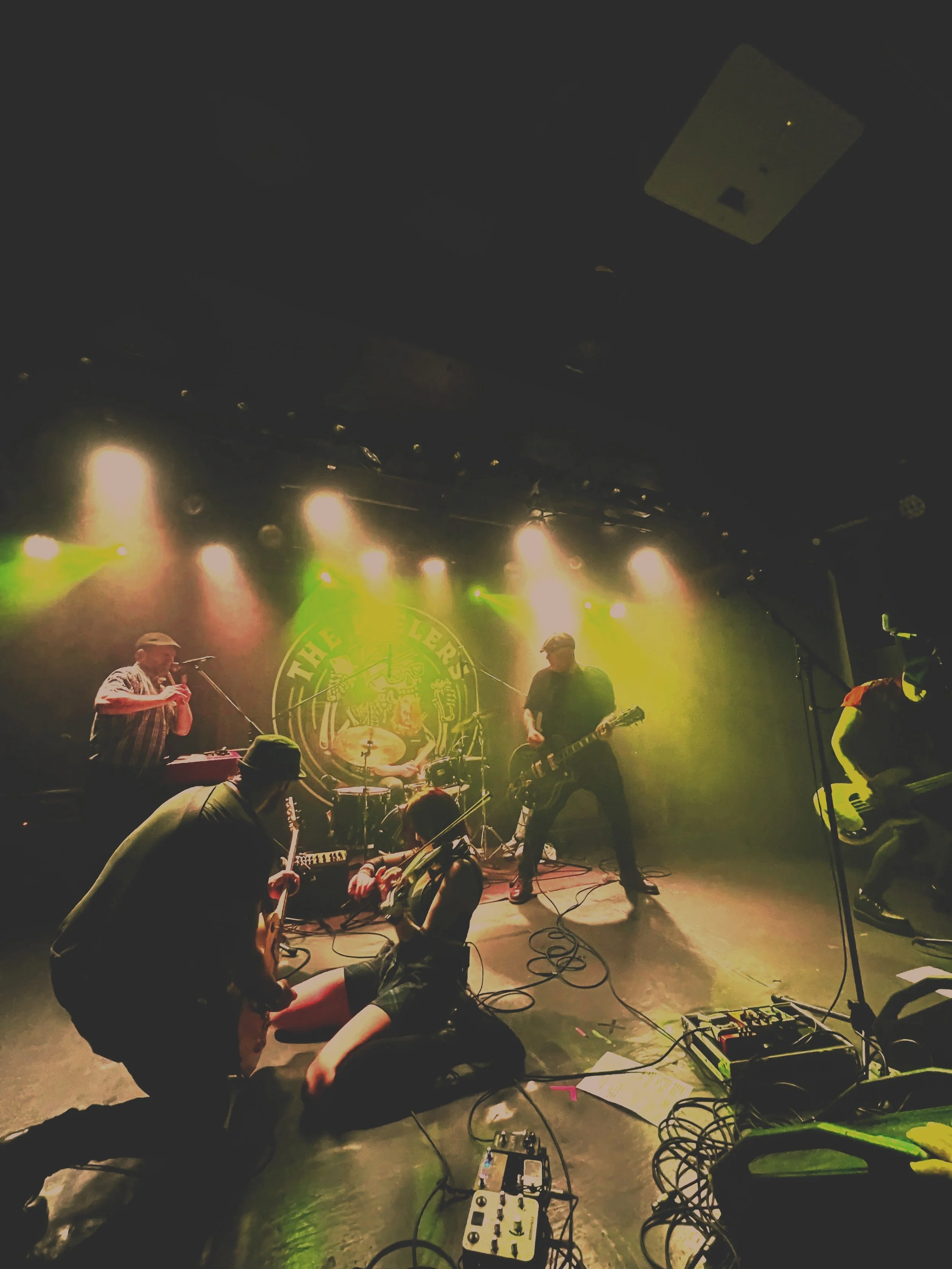 A live band performs on stage with colorful lights, including a singer with a microphone and guitarists, while a person is lying on the floor in front of the stage. Music equipment and cables are visible on the stage.