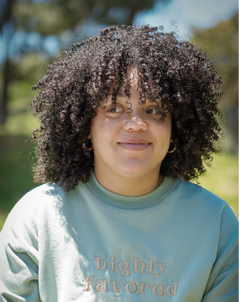 A woman with curly hair wearing a light green sweatshirt that says "highly favored", standing outdoors with trees and a blue sky in the background.