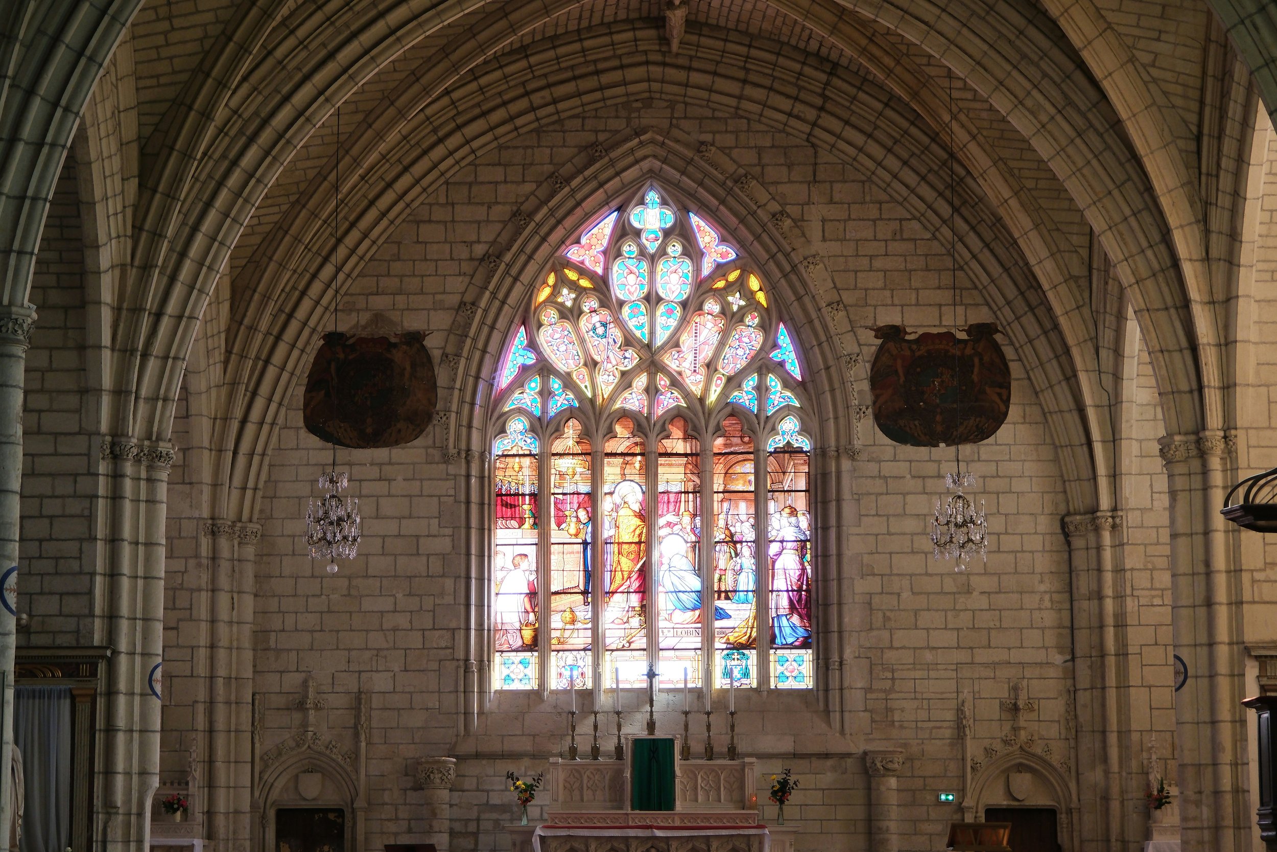 The interior of a church with a large stained glass window depicting a religious scene, stone arches, chandeliers, and flower arrangements at the altar.